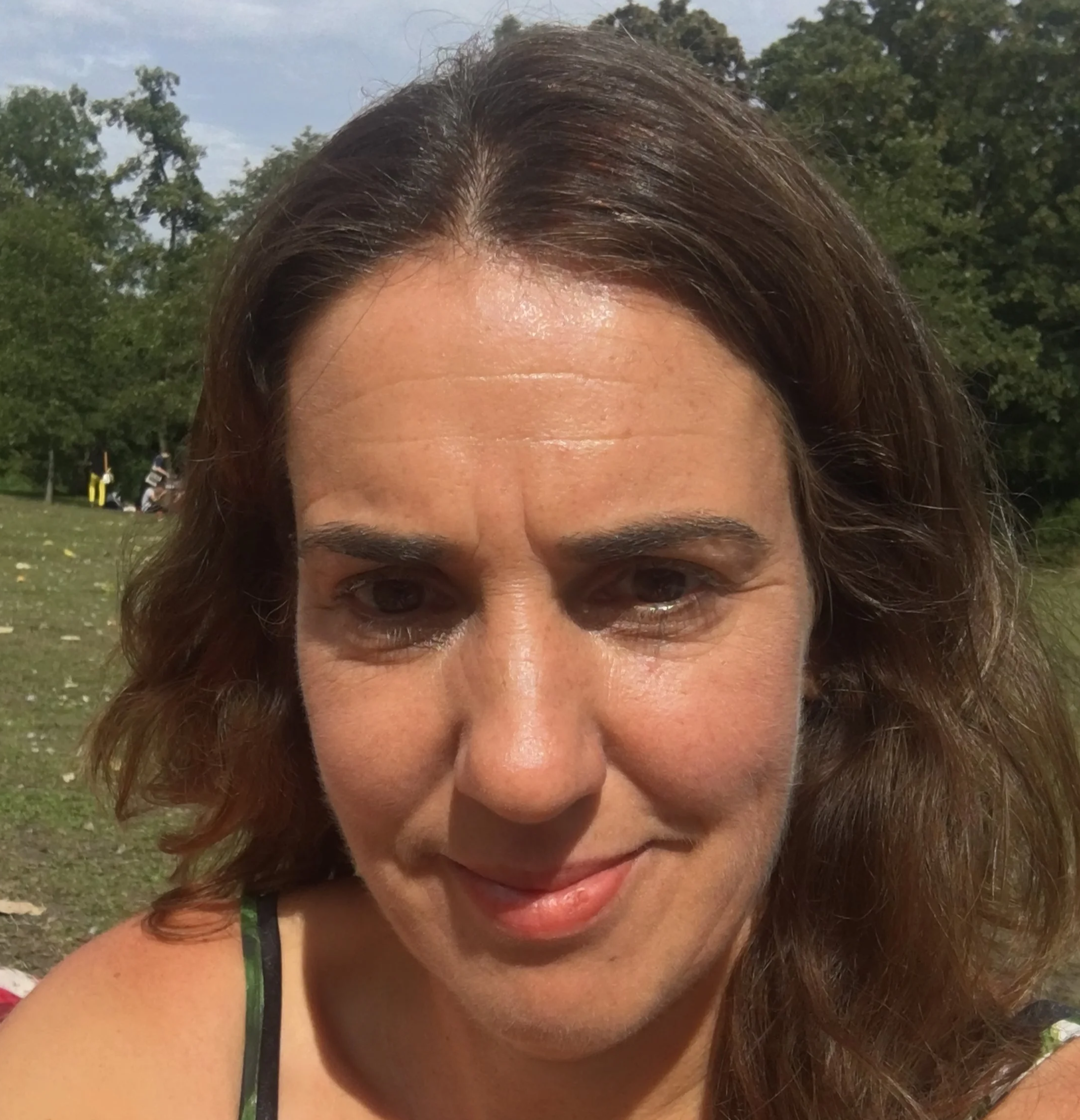 Close-up of a woman with light skin and shoulder-length brown hair outdoors on a sunny day, with trees and people in the background.