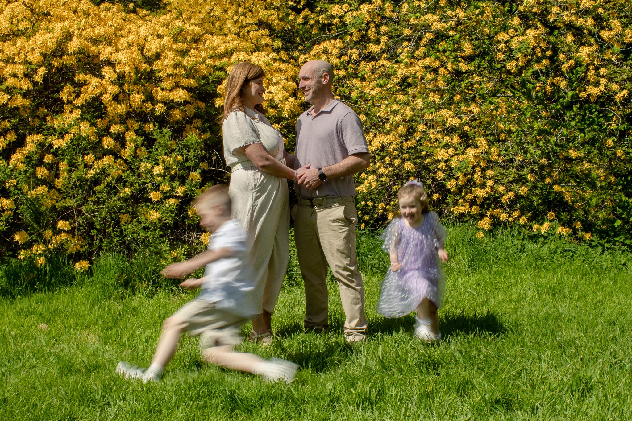 Three children, two girls and a baby, near a calm lake surrounded by trees and greenery, with the older girl smiling and the youngest looking curious.
