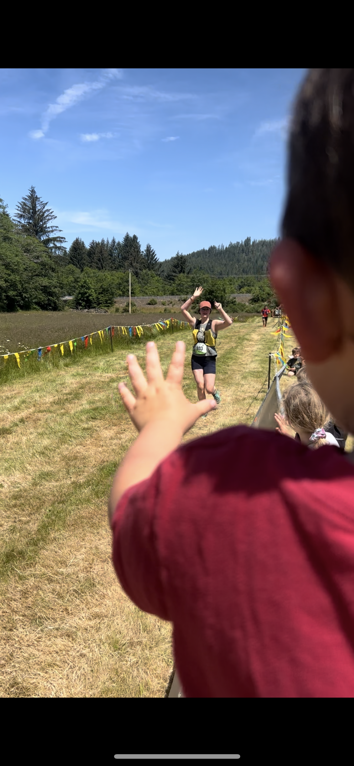 A woman crossing a finish line during a race, with her arms raised in victory, while a child in the foreground reaches out toward her in an outdoor setting on a sunny day.