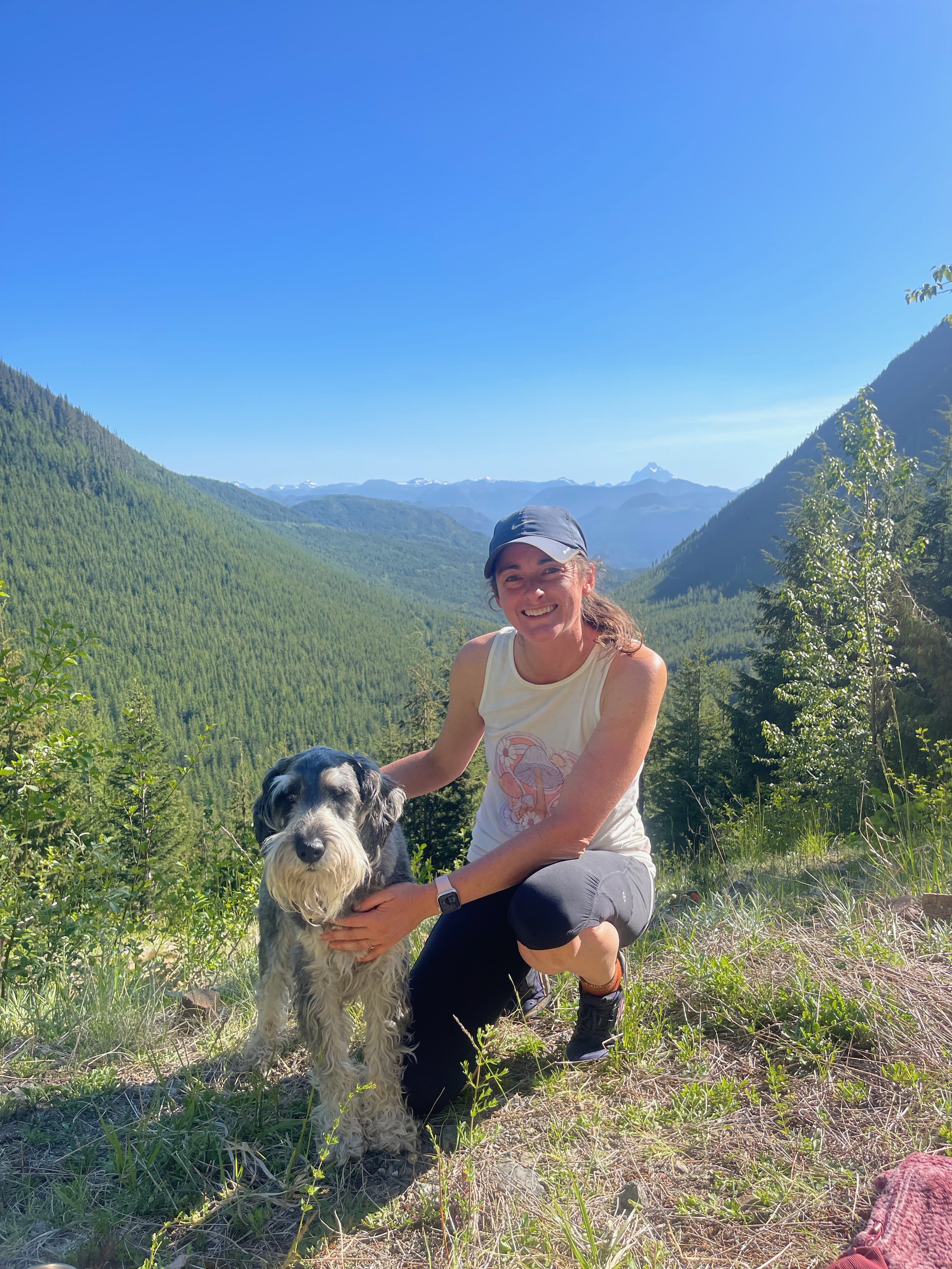 A woman kneeling outdoors with a dog in a scenic mountain landscape under a blue sky.
