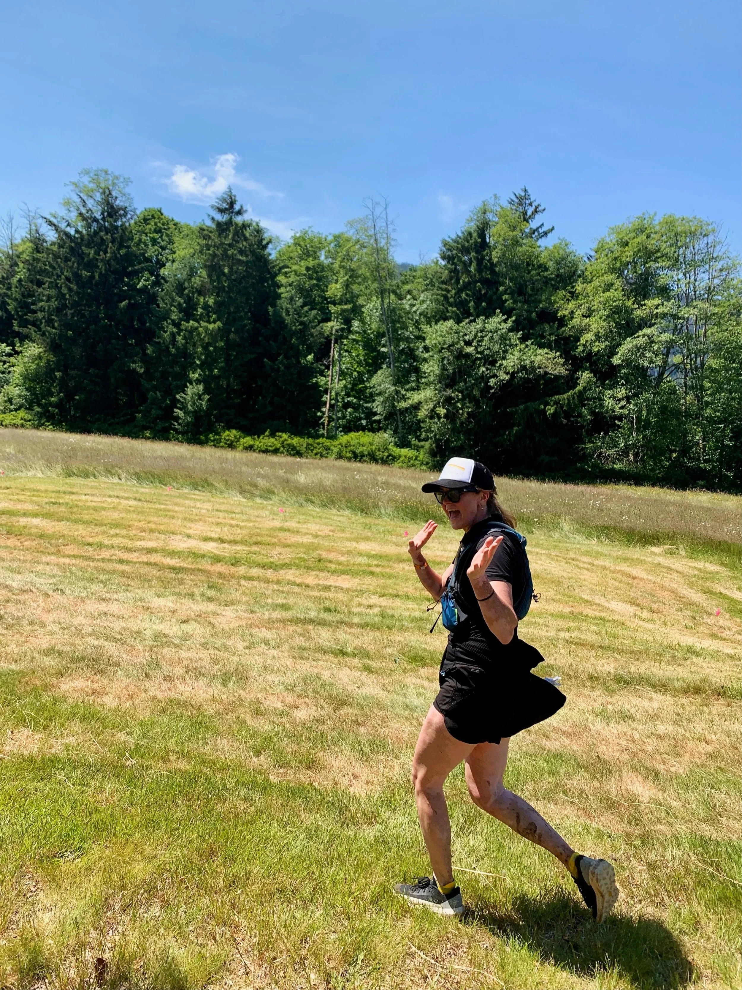 A woman running outdoors on a grassy field, wearing a black cap, sunglasses, black shirt, shorts, and running shoes, surrounded by trees and a blue sky.
