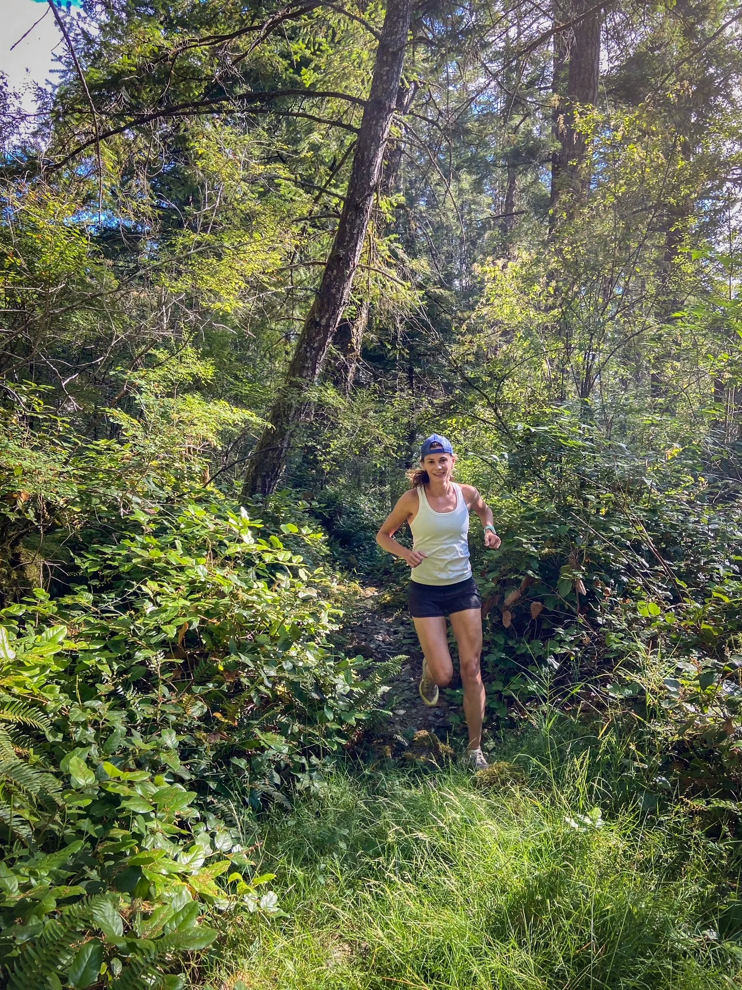 A woman is running on a narrow trail through a lush, green forest with dense foliage, tall trees, and sunlight filtering through the leaves.