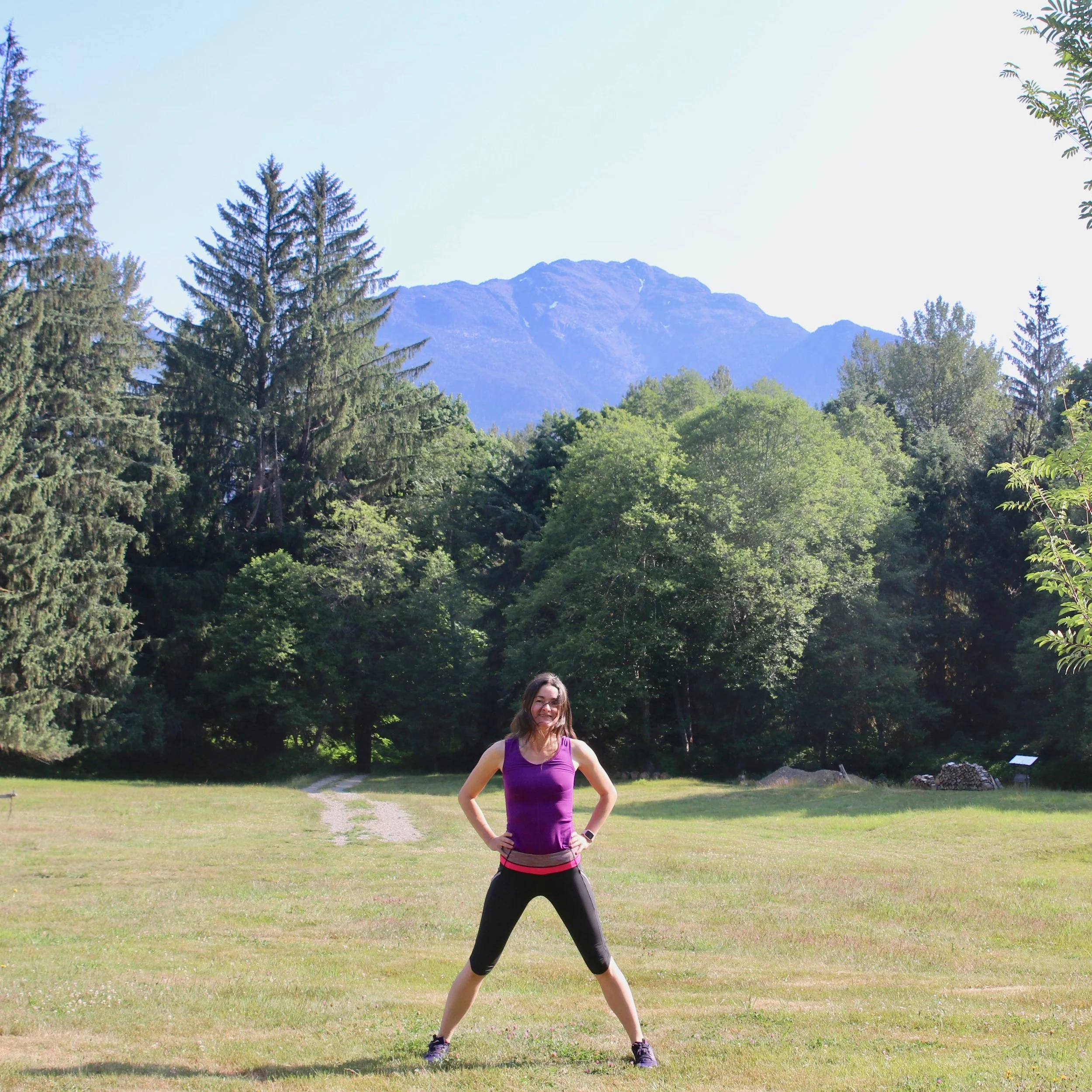 A woman standing outdoors in a grassy field with her hands on her hips, wearing a purple tank top and black leggings, with a backdrop of trees and mountains under a clear sky.