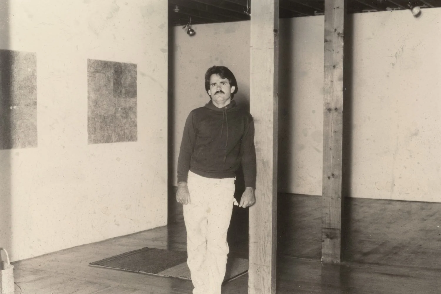 A man with a mustache and dark hair standing indoors near a wooden partition, wearing a dark hoodie and light-colored pants.