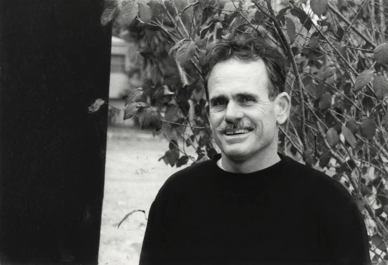 Black and white photo of a smiling man with a mustache, wearing a dark sweater, standing outdoors next to leafy bushes.