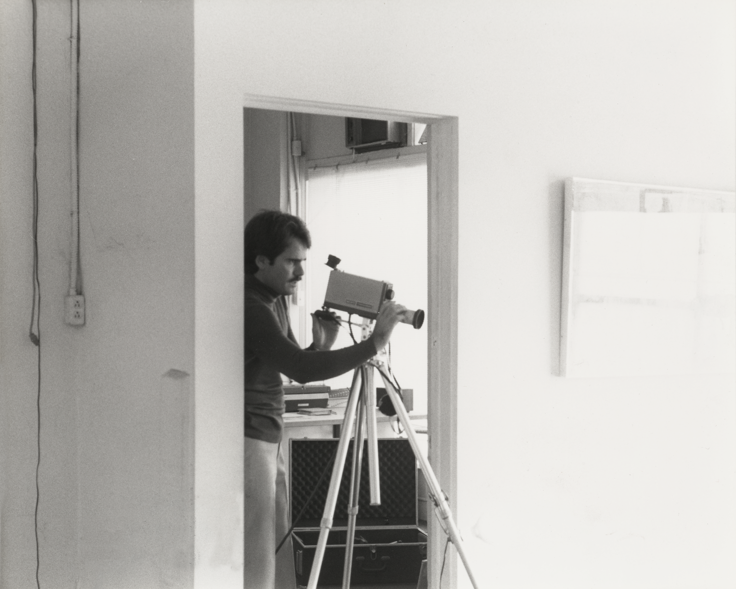 A man operating a vintage movie camera on a tripod inside a room, with white walls and some artwork hanging, as seen through a doorway.