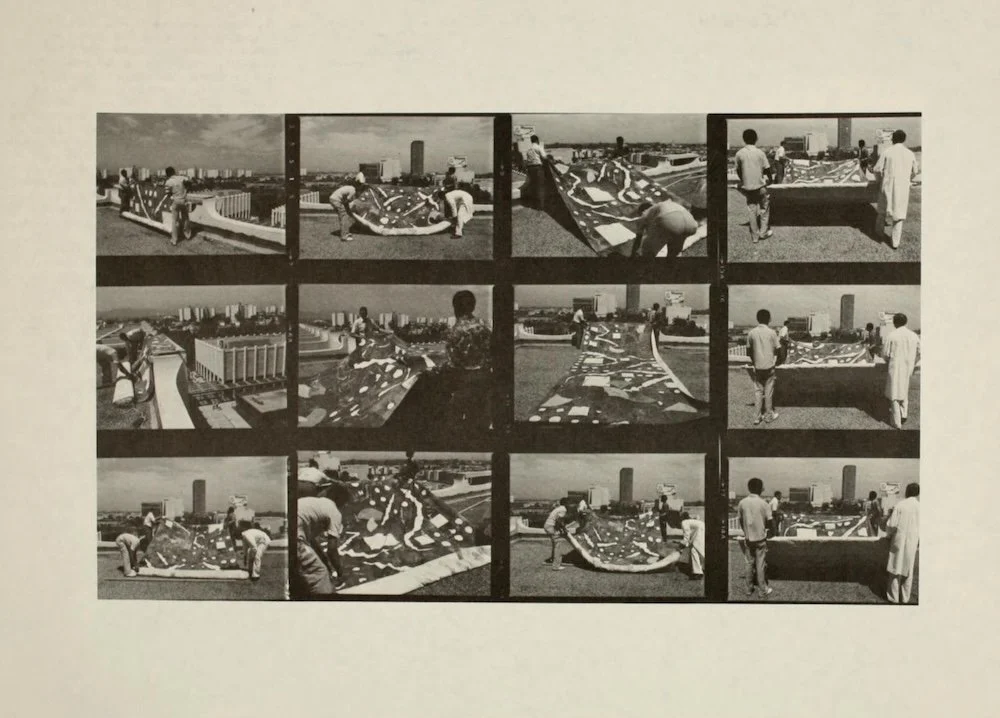 Black and white collage of people assembling a large model or map on a rooftop with city skyline in the background.