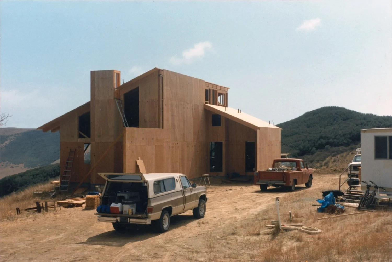 Wooden house under construction with various vehicles and construction materials in the foreground, hills in the background.
