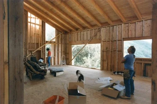 Two women and a dog in an unfinished wooden house under construction, with large open windows and construction materials scattered around.