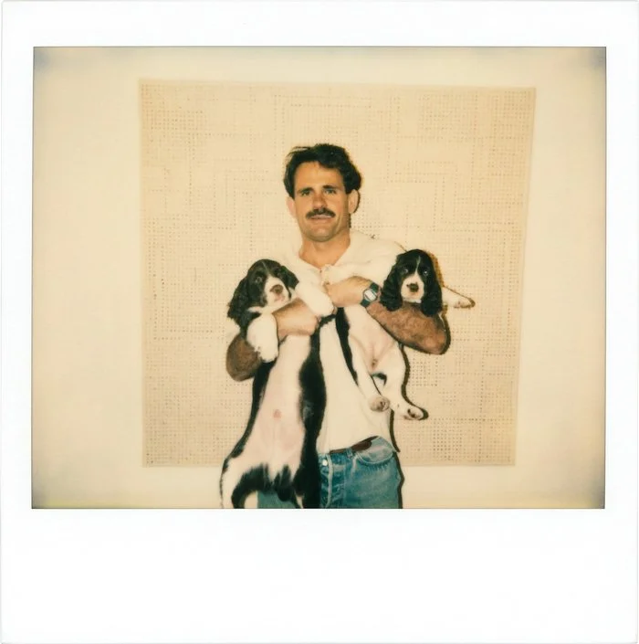 A man with dark hair, mustache, wearing a white shirt and jeans, holding two puppies with black and white fur, standing in front of a pegboard wall.