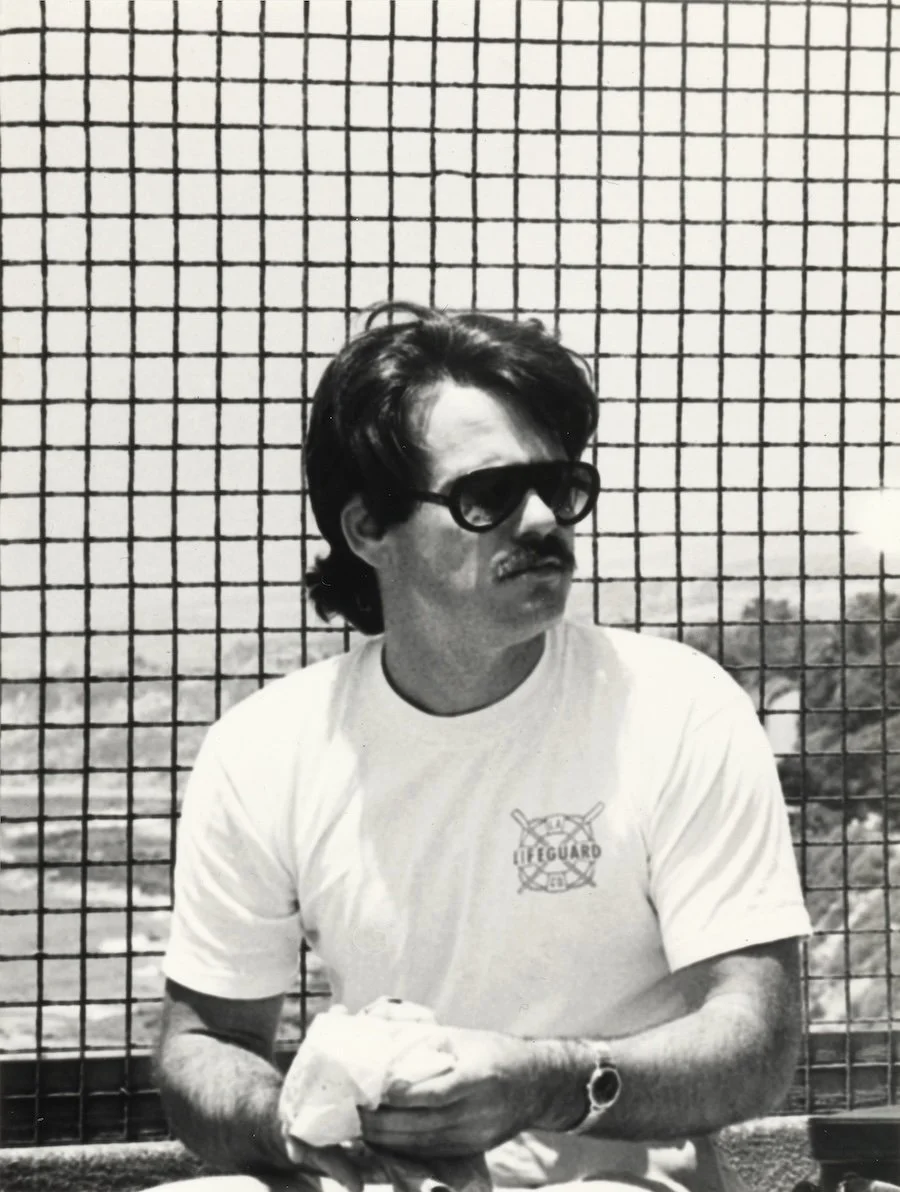 Man in sunglasses and a lifeguard T-shirt sitting outdoors with a fence behind him, holding an object wrapped in cloth.