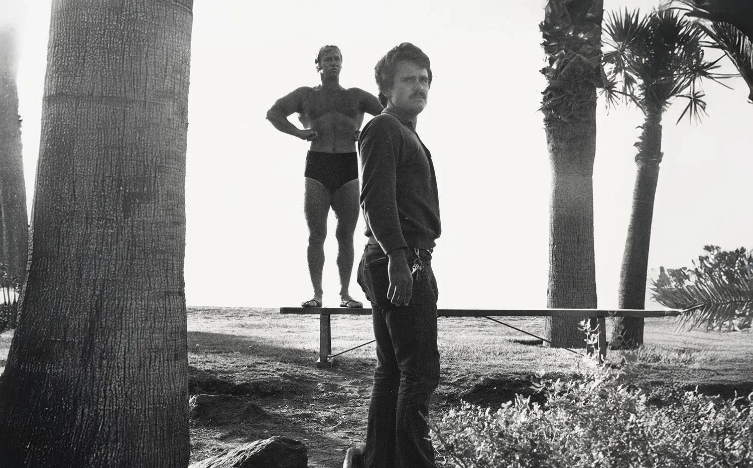 A black and white photo of a man standing on the ground with a surfboard balanced on a thin support behind him, while a woman stands behind it on the support with her hands on her hips, near palm trees in the background.