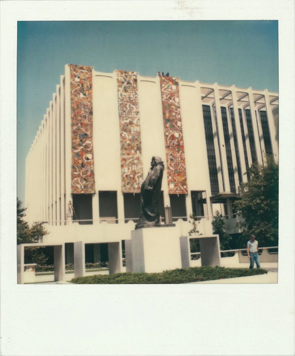 A large building with vertical white beams, colorful mosaic panels, and a statue of a robed figure in front.