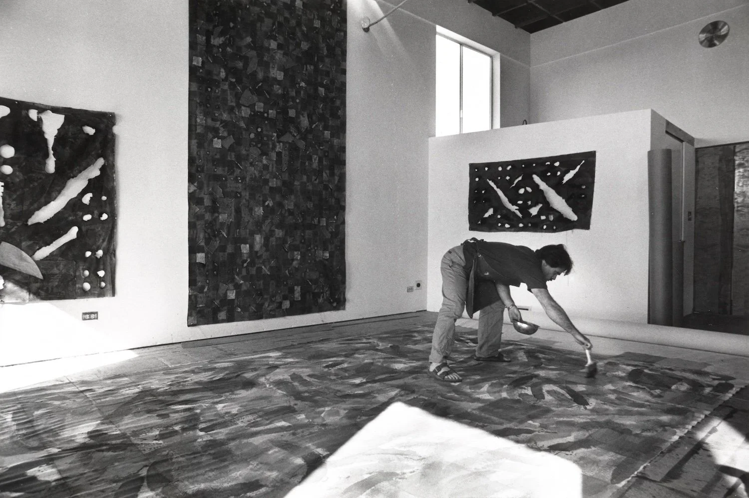 An artist is working on a large abstract painting on the floor of an art gallery with two large abstract paintings hanging on the walls behind them.