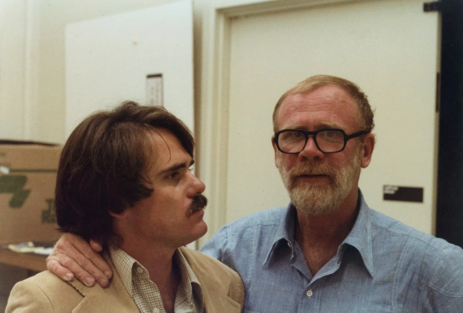 Two men in an indoor setting; one with dark hair and a mustache, wearing a beige jacket, and the other with a beard, glasses, and light blue shirt, standing close together with the man in the blue shirt resting his hand on the other's shoulder.
