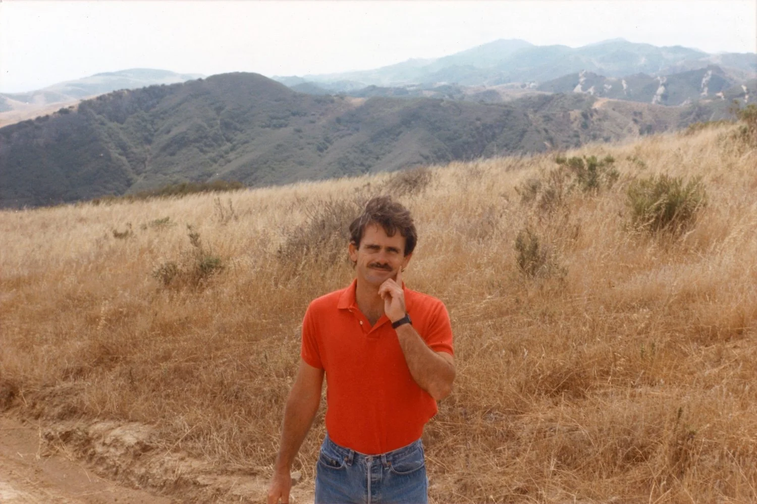 A man with dark hair, mustache, wearing a red polo shirt, blue jeans, and a black wristwatch, stands outdoors in a dry, grassy field with rolling hills and mountains in the background.