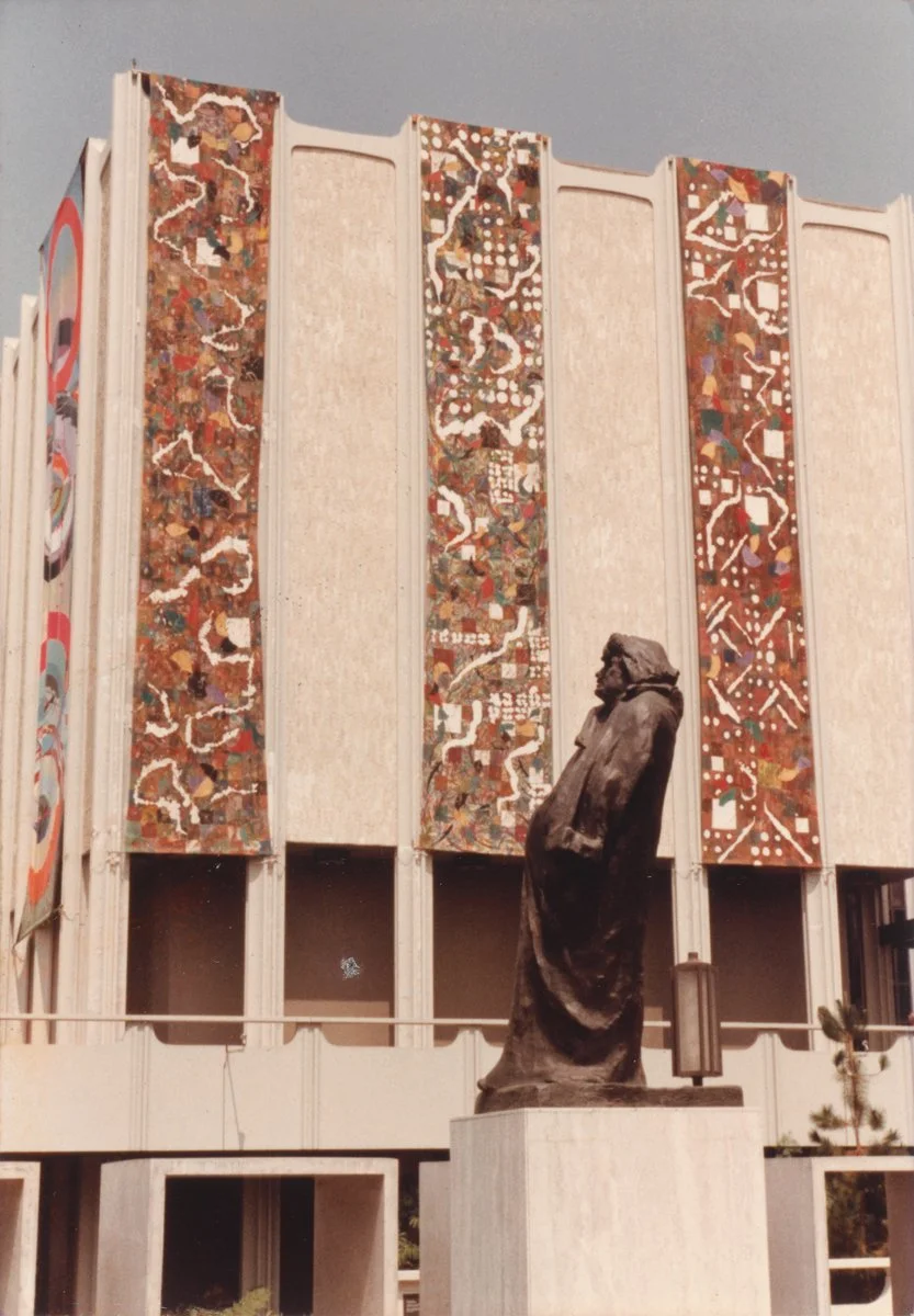 A bronze statue of a man stands in front of a modern building with colorful, patterned vertical panels. The statue depicts a man with a beard, dressed in a coat, looking upward.