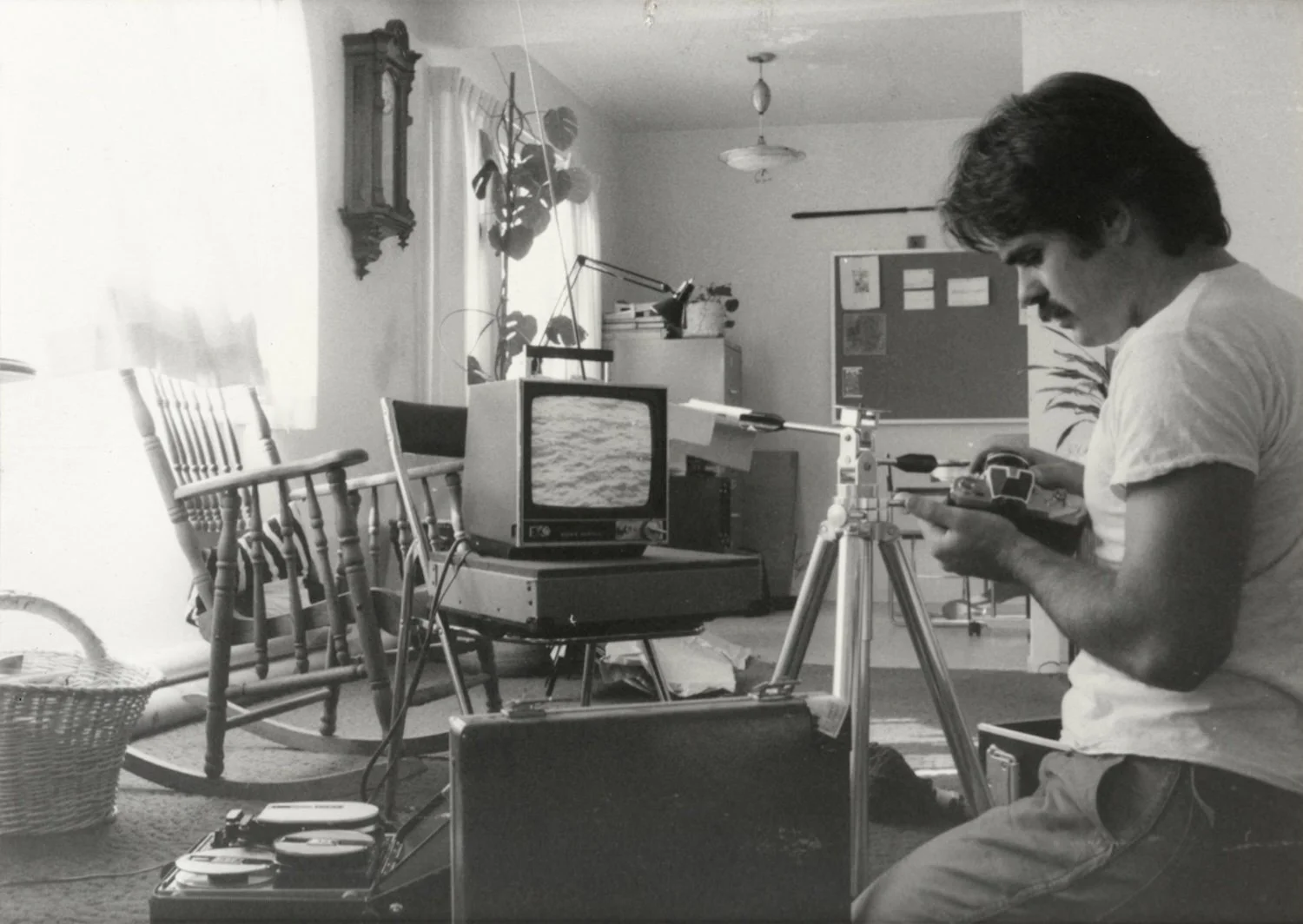 A man with dark hair and a mustache is sitting on the floor in a room, operating a camera mounted on a tripod while looking at a remote control. Behind him is a vintage television displaying a body of water, and toy train tracks are laid on the floor nearby. The room has chairs, a wicker basket, a doorway with blinds, and a wall-mounted clock.