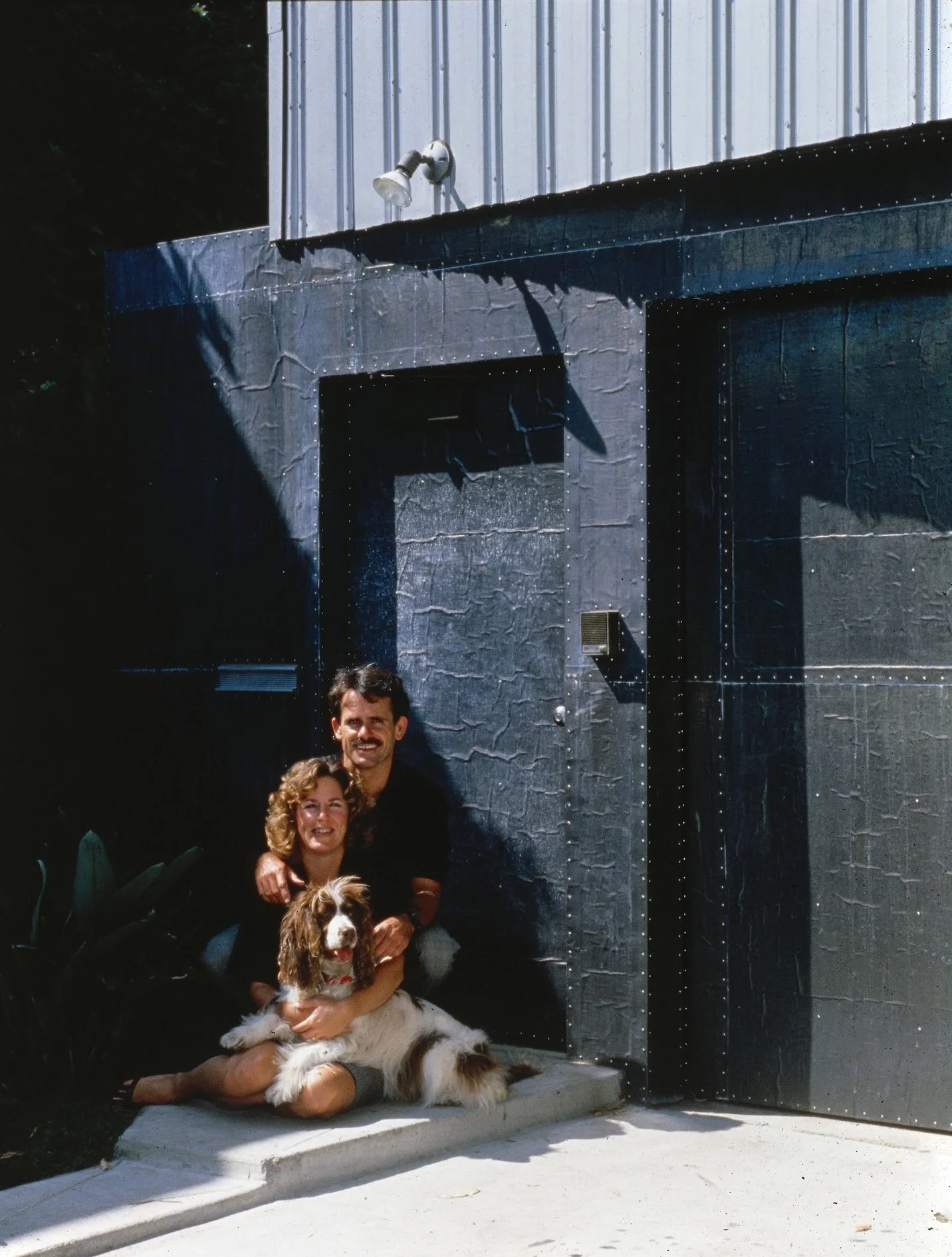 A happy couple sitting on the sidewalk outside a black building, holding their dog.
