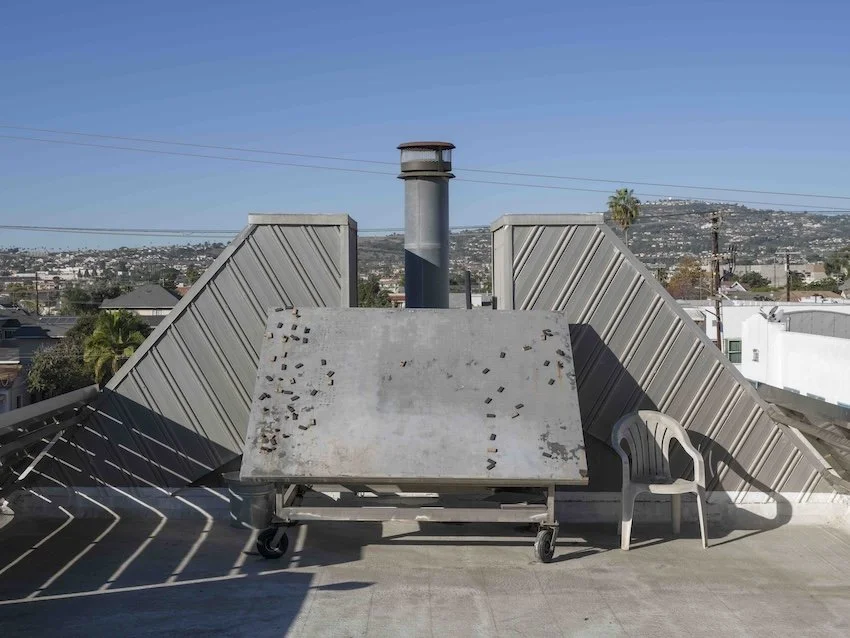 Rooftop with a metal tabletop, a plastic chair, and a chimney, overlooking a city and hills in the distance.