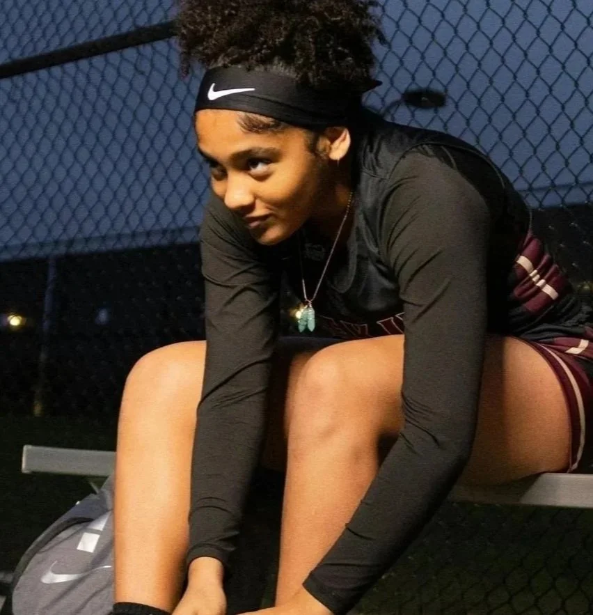 A young female athlete with curly hair in a black Nike headband, wearing a black sports long-sleeve shirt and maroon sports shorts, sitting on a bench at a sports court during dusk.