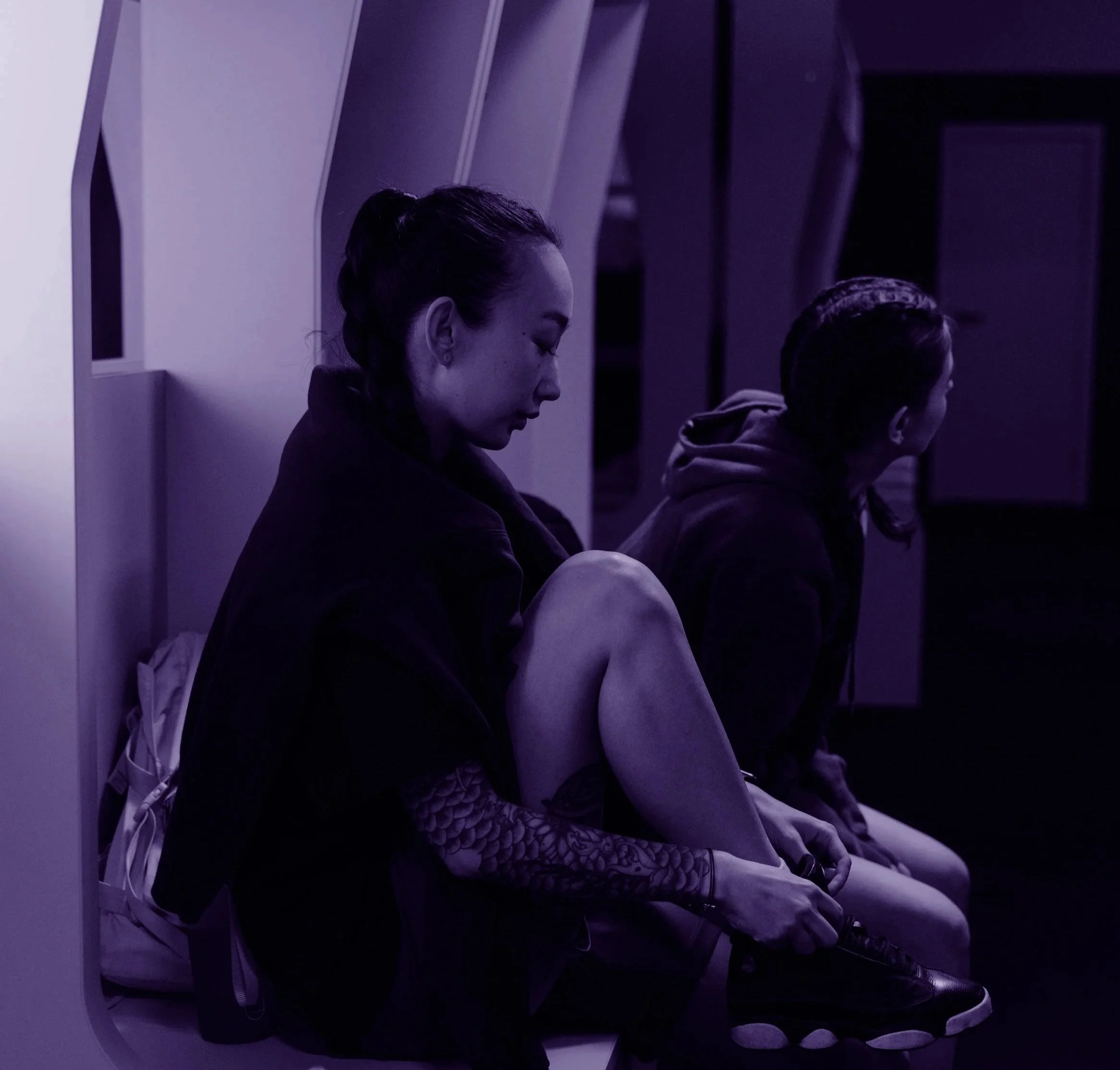 Two young women sitting on a bench in a dimly lit space, one with her eyes closed and head bowed, the other looking away, both with braided hair, dark clothing, and sneakers, in a contemplative posture.