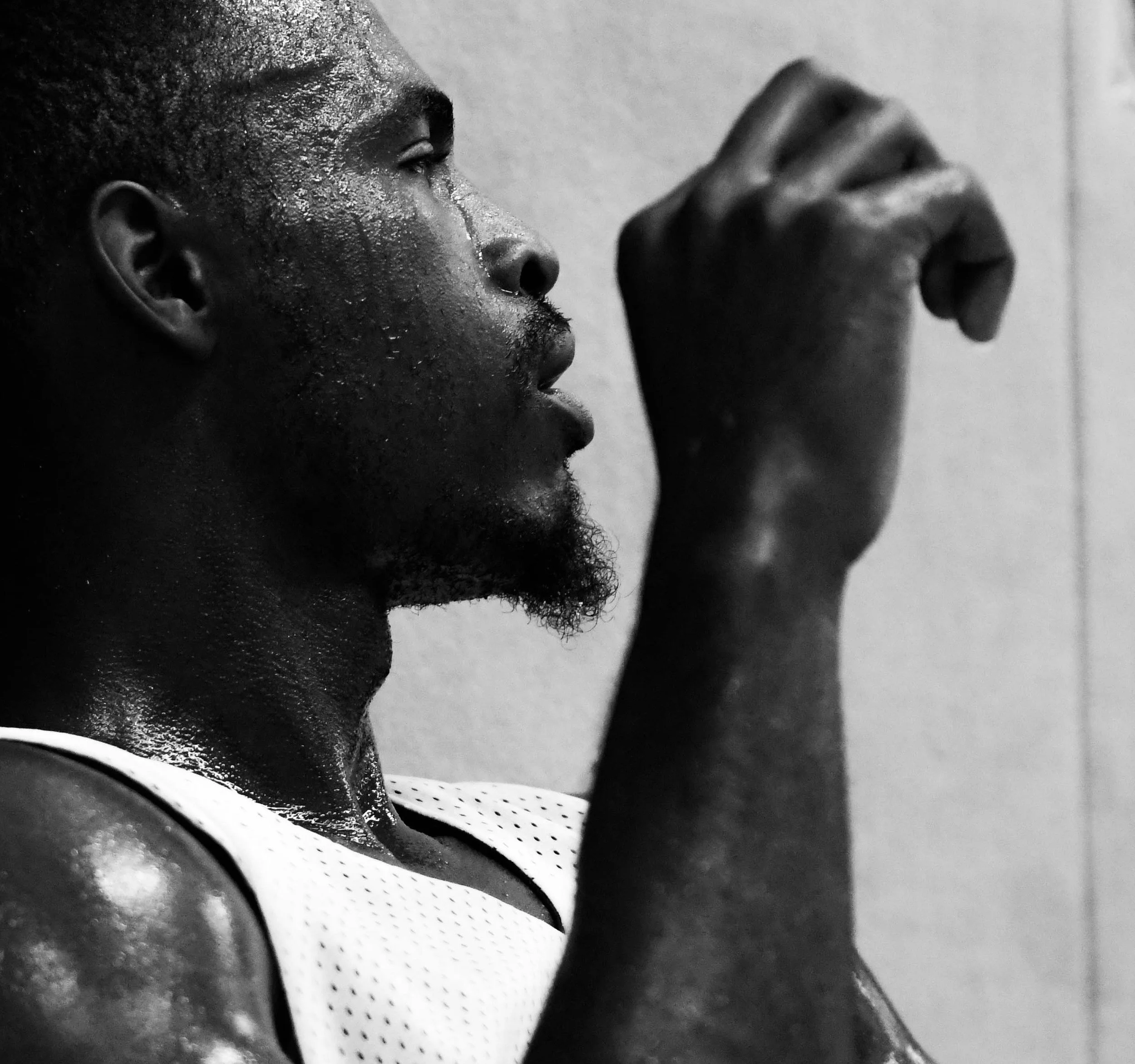 A close-up black and white photo of an athletic man with sweat on his face, gazing into the distance, wearing sports attire, with one hand raised near his face.