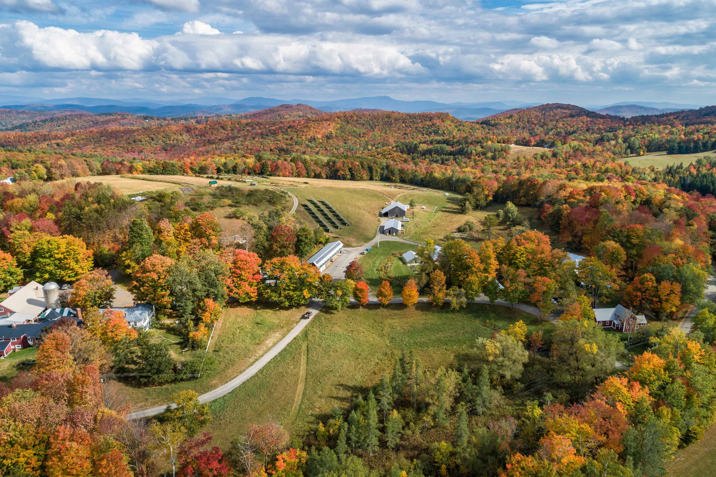 Aerial view of a rural area during fall with colorful autumn trees, green fields, and scattered houses, with distant rolling hills under a partly cloudy sky.