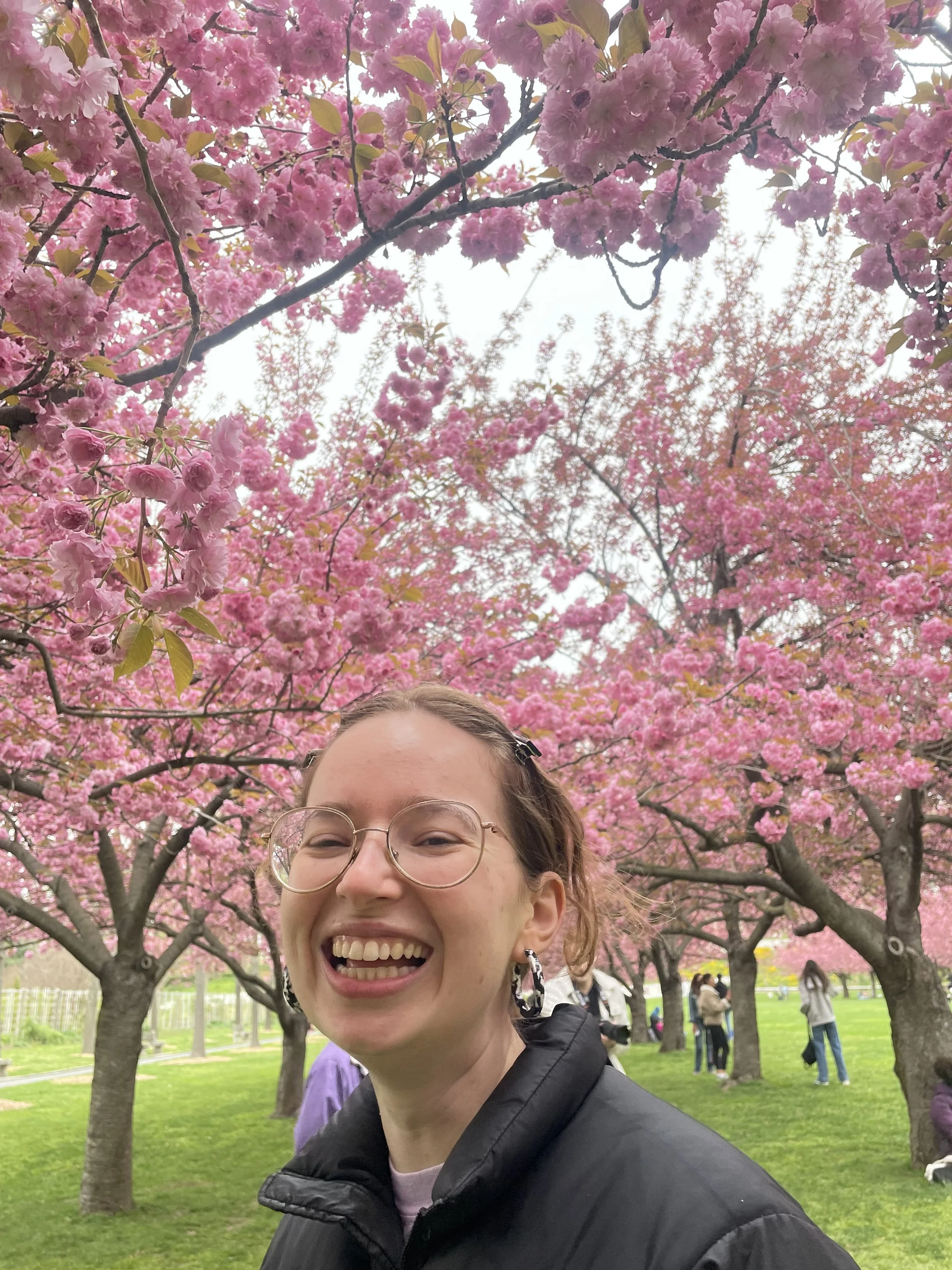 A young woman with glasses and earrings smiling in front of blooming pink cherry blossom trees at a park on a cloudy day.