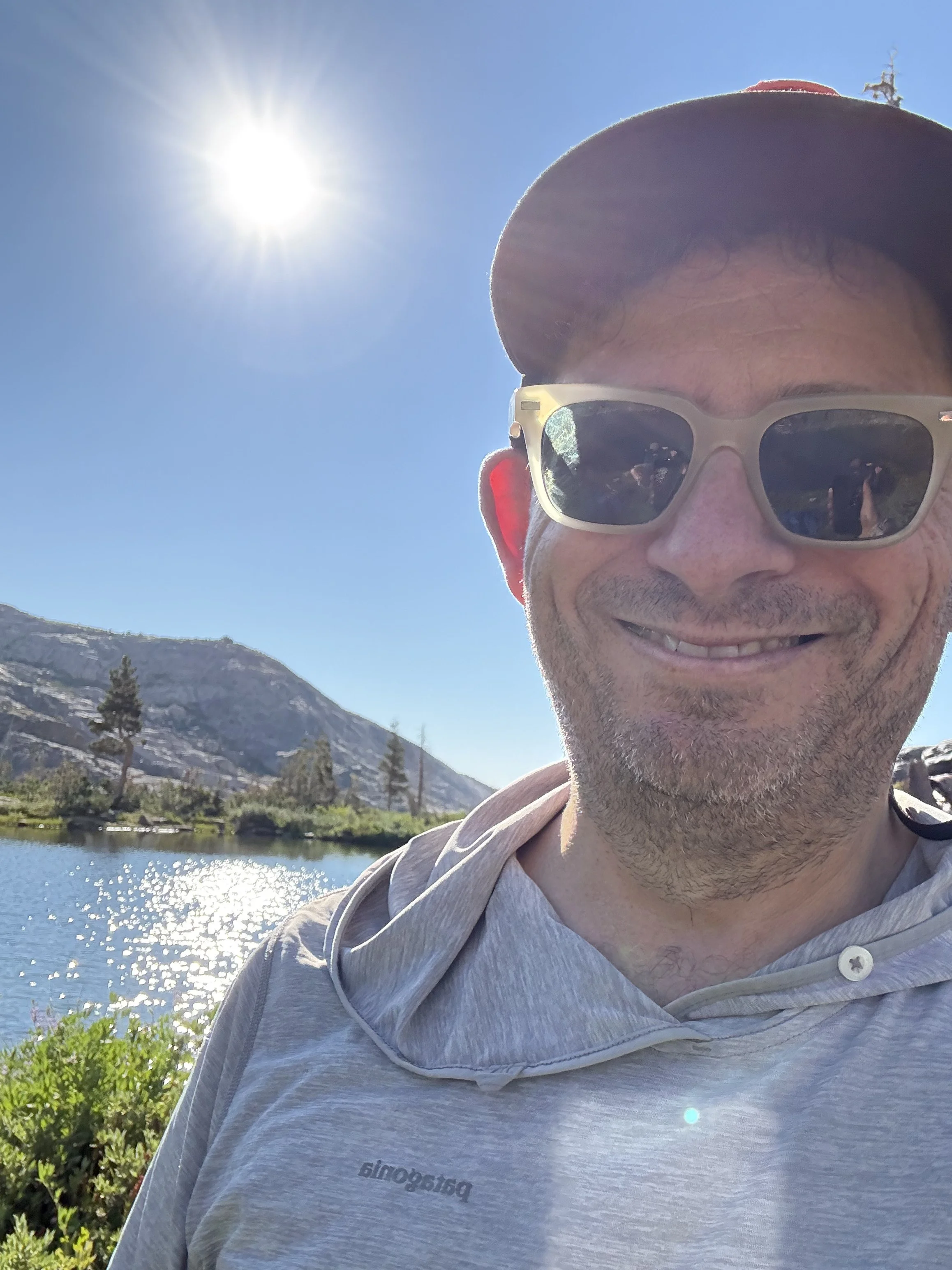 A man outdoors near a lake with mountains in the background, wearing sunglasses and a baseball cap, smiling on a sunny day.