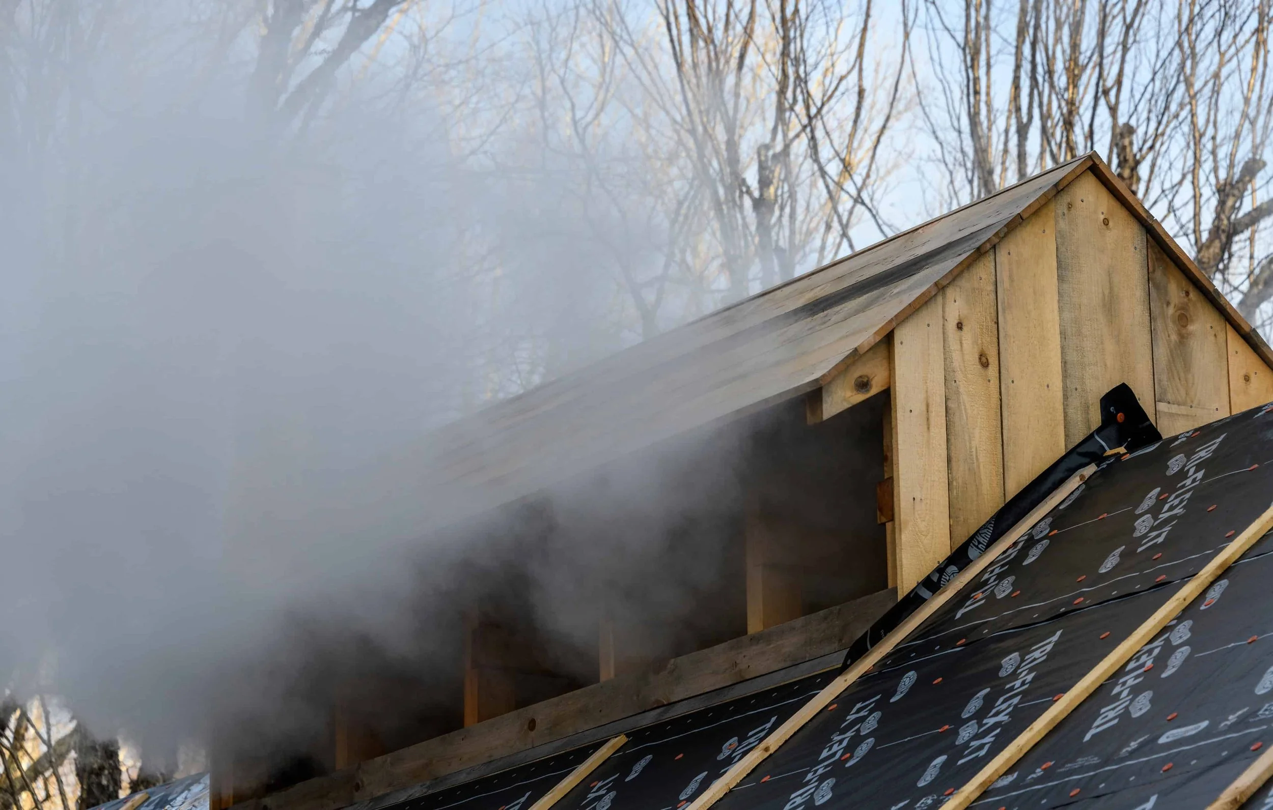 Construction of a small wooden sugaring shed with a sloped roof, surrounded by trees with bare branches, and a steam cloud rising out of the roof for maple syrup production.