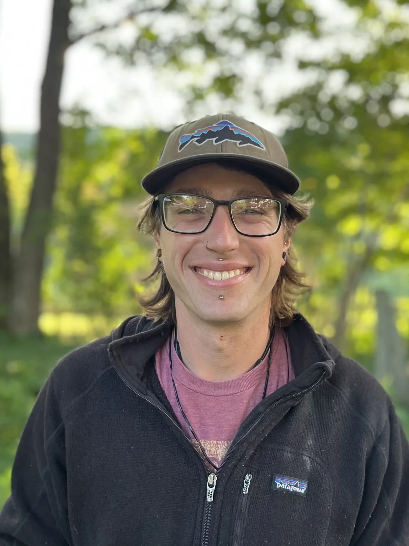 A person smiling outdoors wearing glasses, a baseball cap with a mountain logo, a pink shirt, and a black Patagonia jacket, with leafy trees in the background.