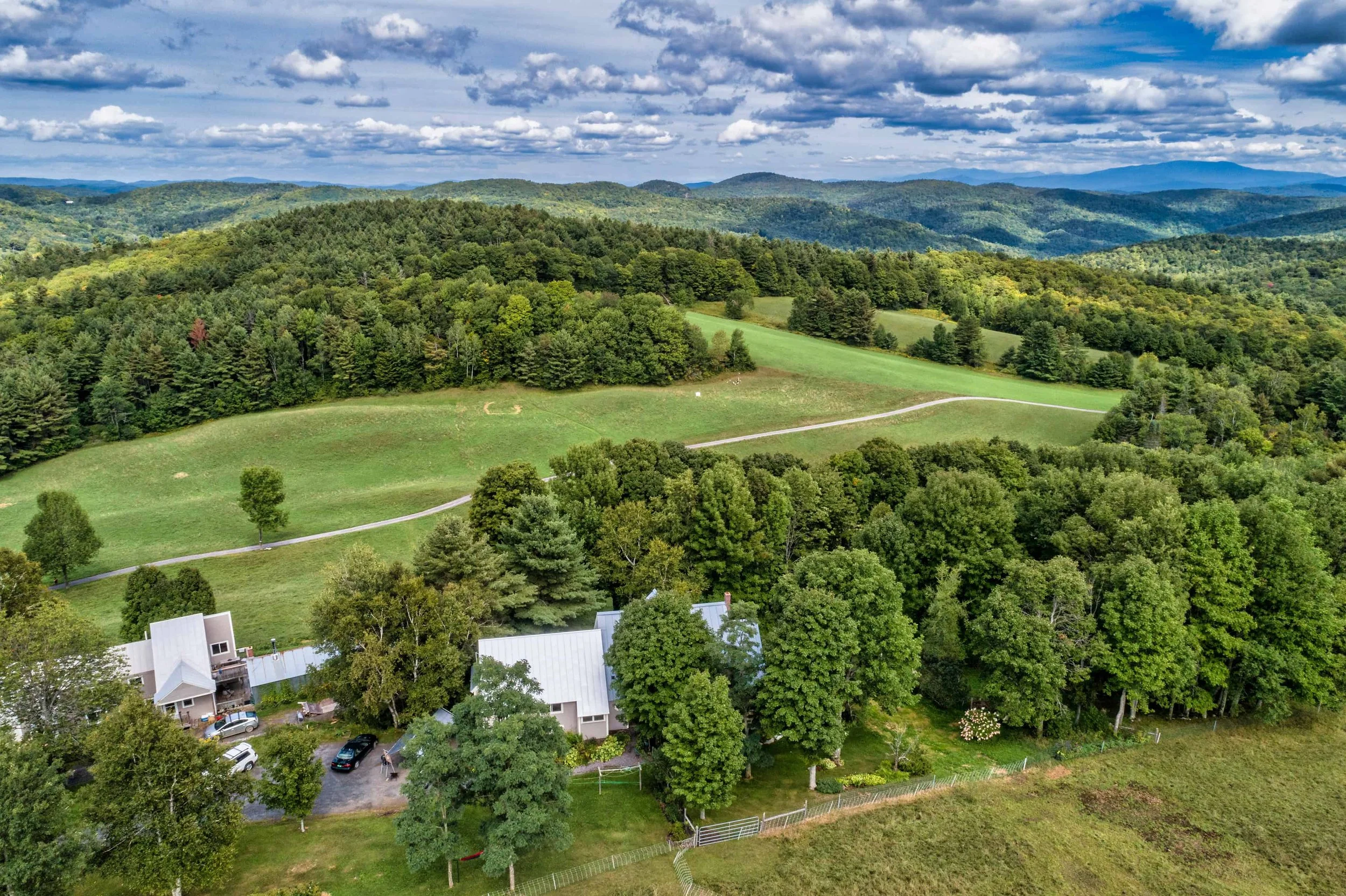 A scenic view of green rolling hills and dense forests with a small residential area at the bottom, under a partly cloudy sky.
