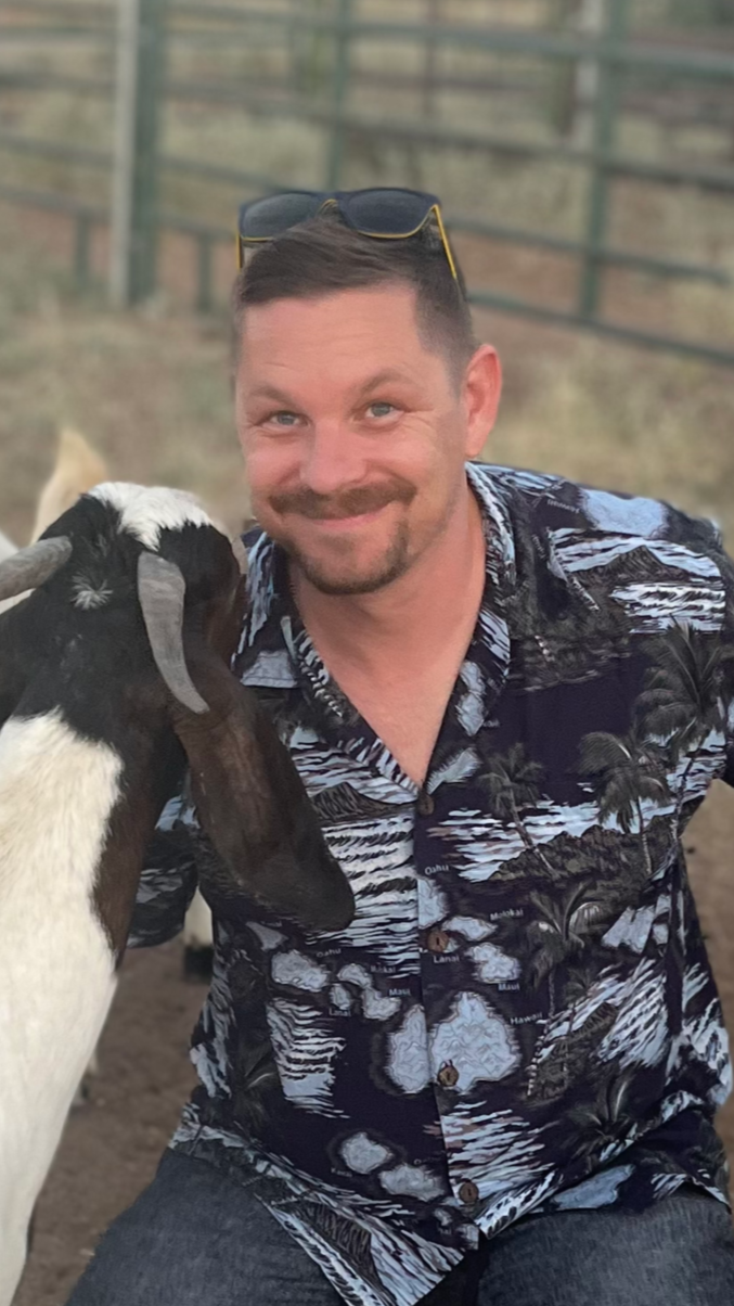 A man with sunglasses resting on his head smiling as a goat nuzzles his face, outdoors with a fence in the background.