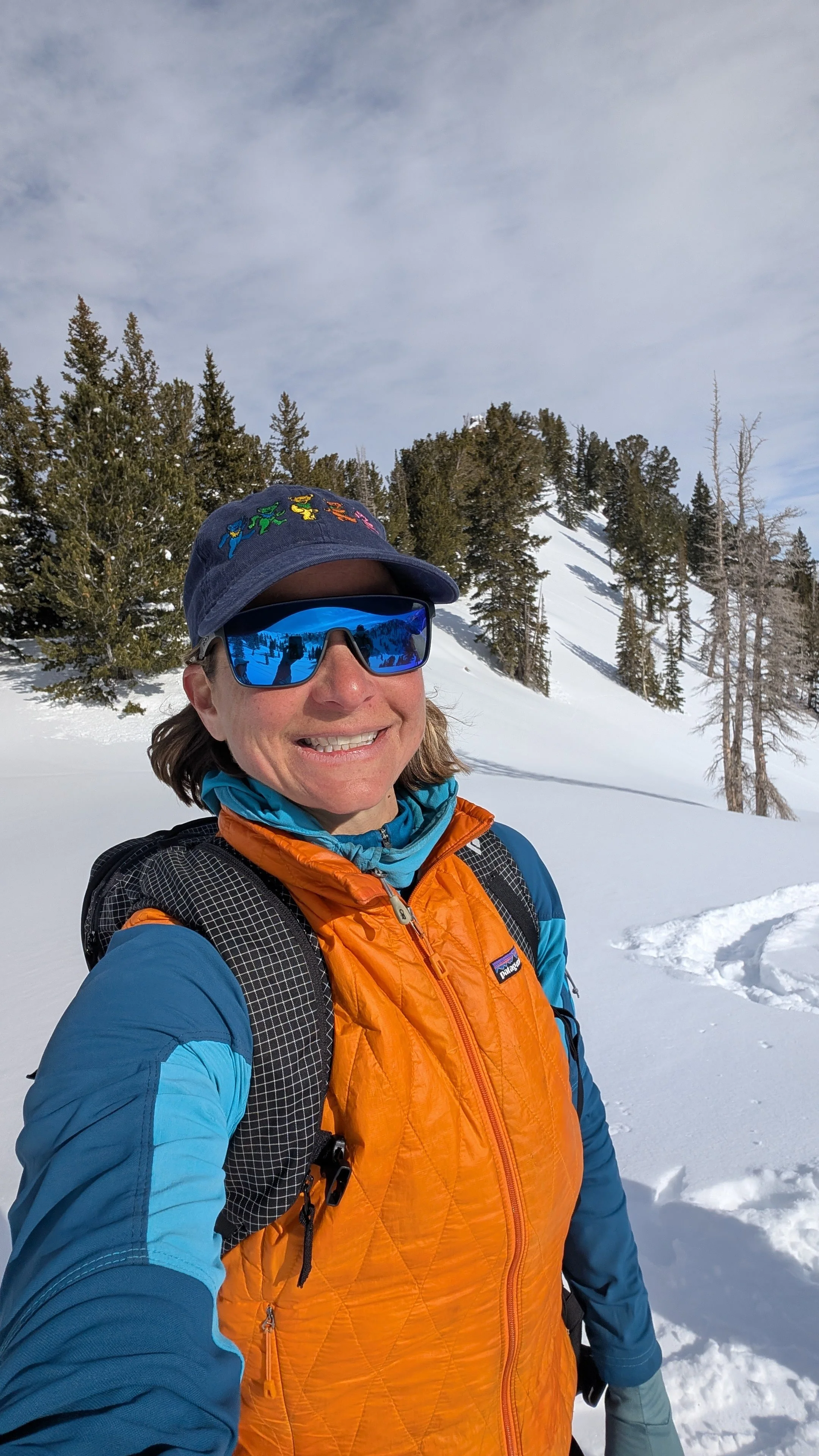 Woman wearing sunglasses, a blue hat with colorful frogs, and an orange and blue jacket, taking a selfie in a snowy mountain landscape with trees and a partly cloudy sky.