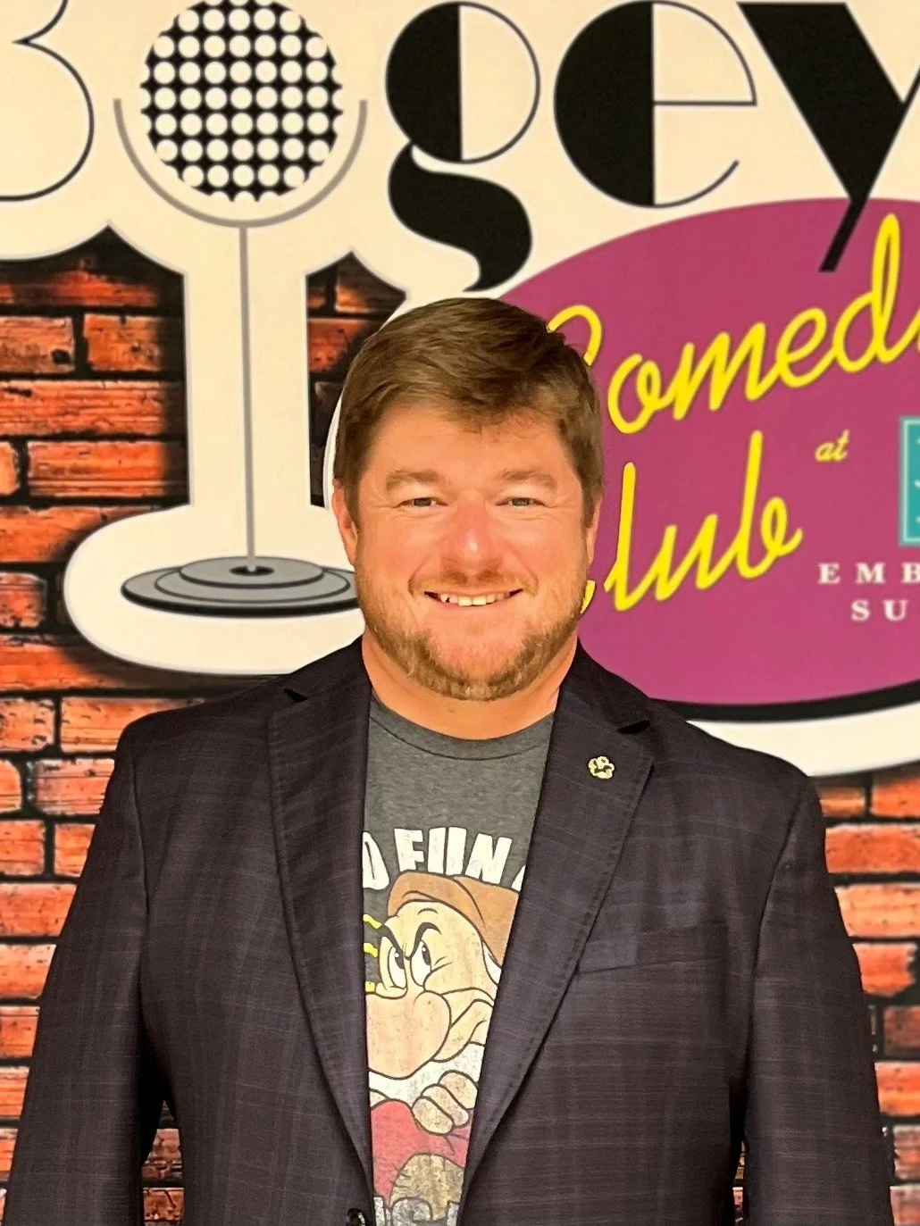 A man with brown hair and a beard smiling in front of a comedy sign with a brick wall background, wearing a black blazer and a graphic t-shirt.