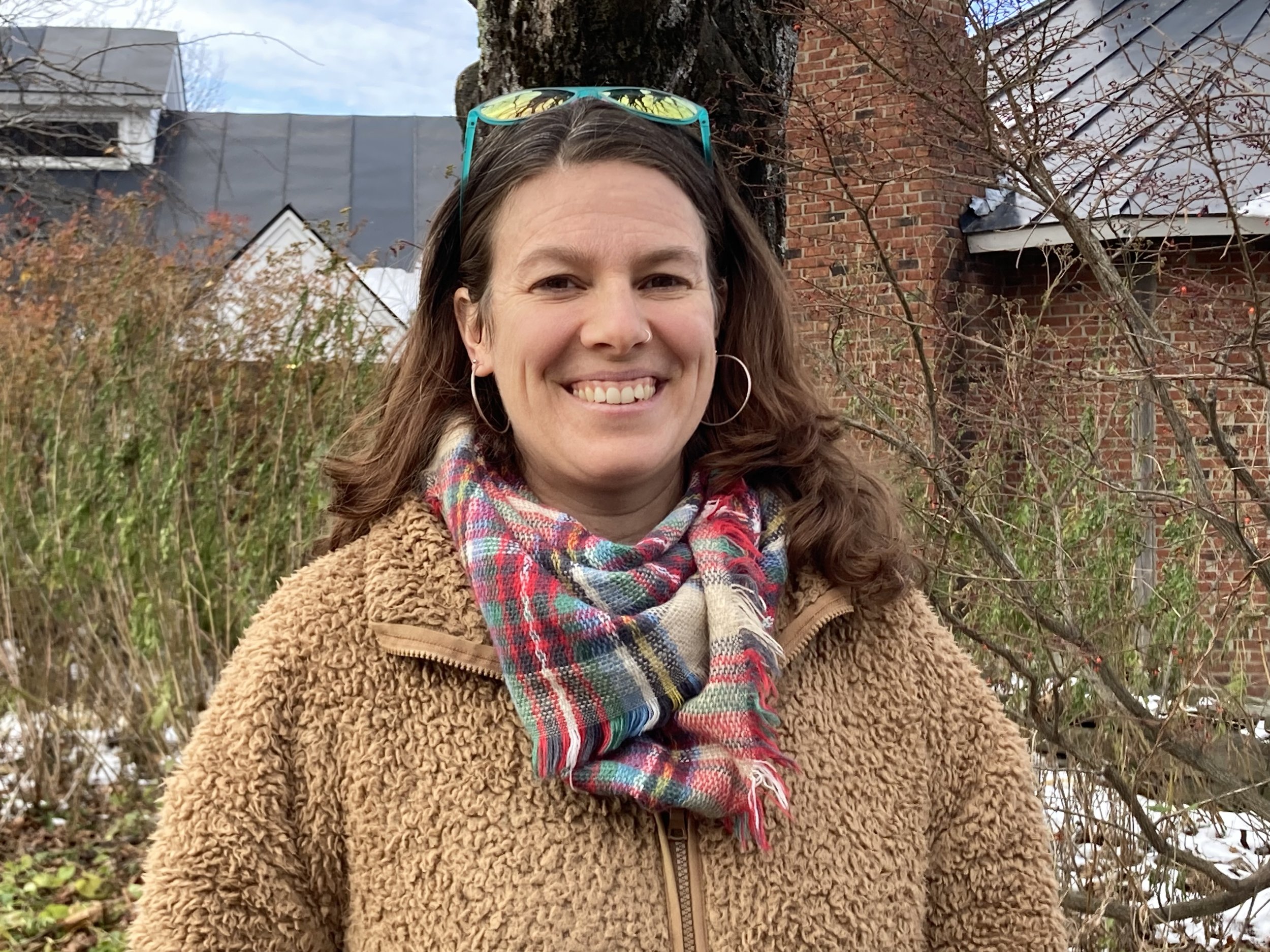 A woman with brown hair, smiling, wearing hoop earrings, a colorful plaid scarf, and a tan, fuzzy zip-up jacket standing outdoors near leafless trees and brick buildings.