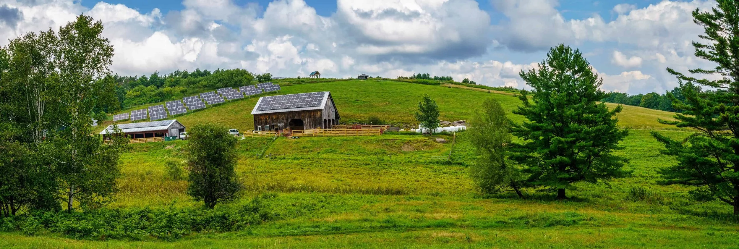 Green hilly farmland with a barn and house covered in solar panels, trees, and a cloudy sky.