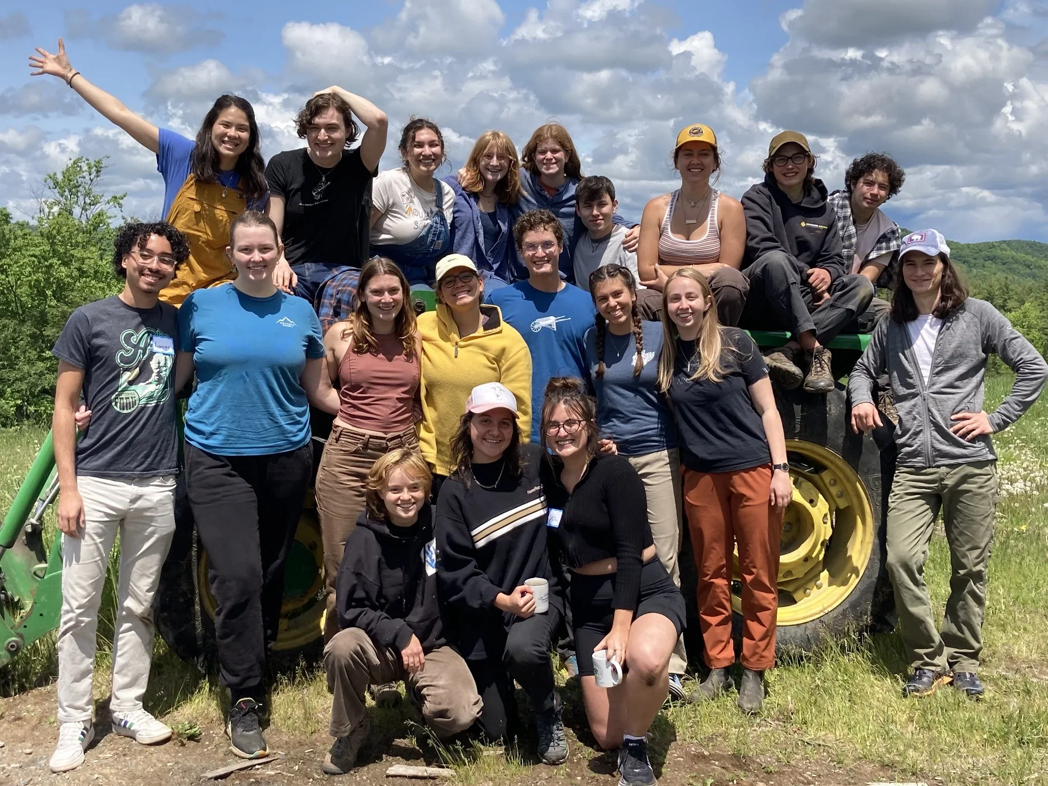 Group of young adults and teenagers posing outdoors on a tractor in a green field with trees and clouds in the sky