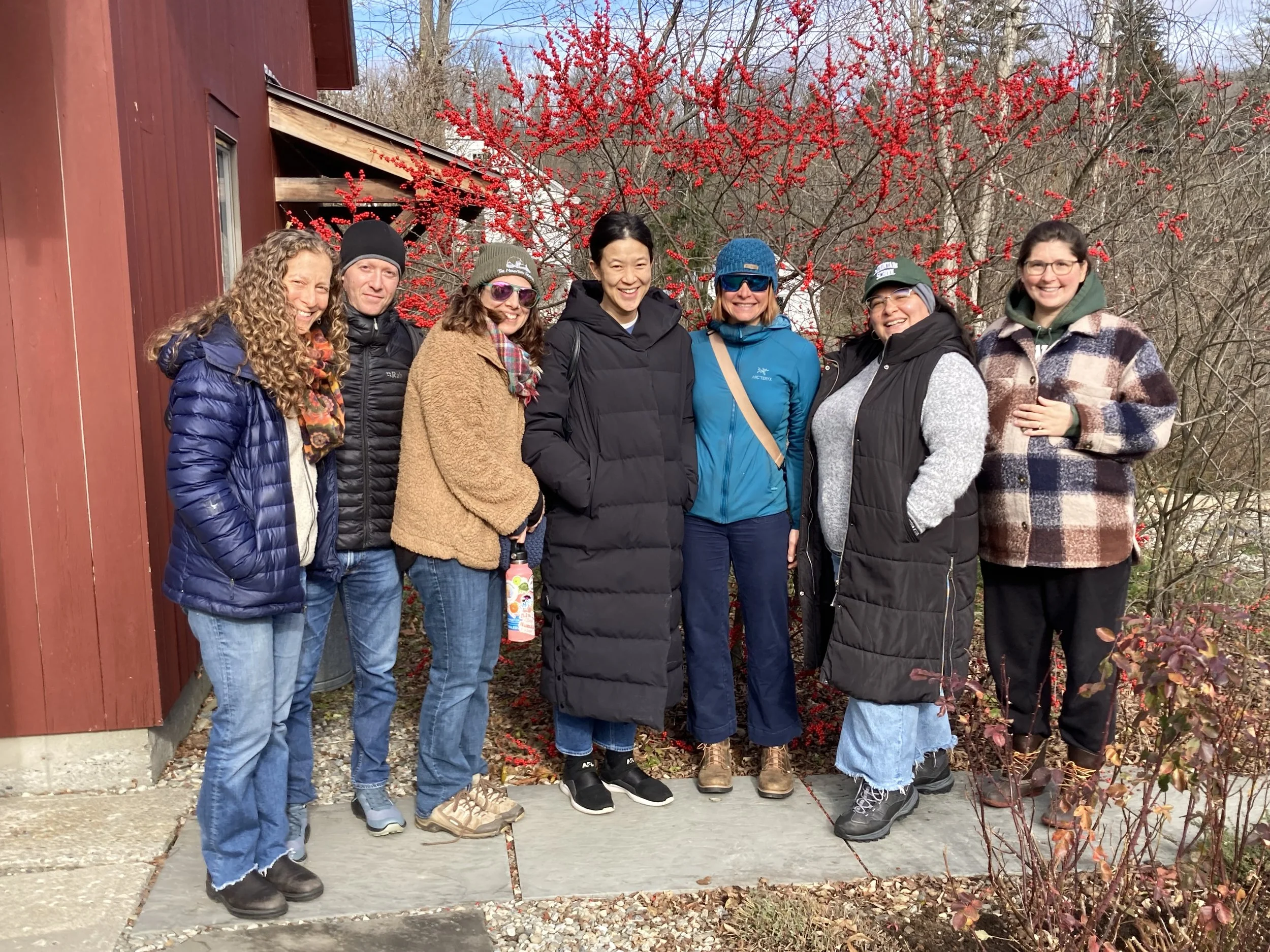 Group of eight women standing outside in front of a red building and red bush, dressed in warm clothing, smiling, with trees in the background.