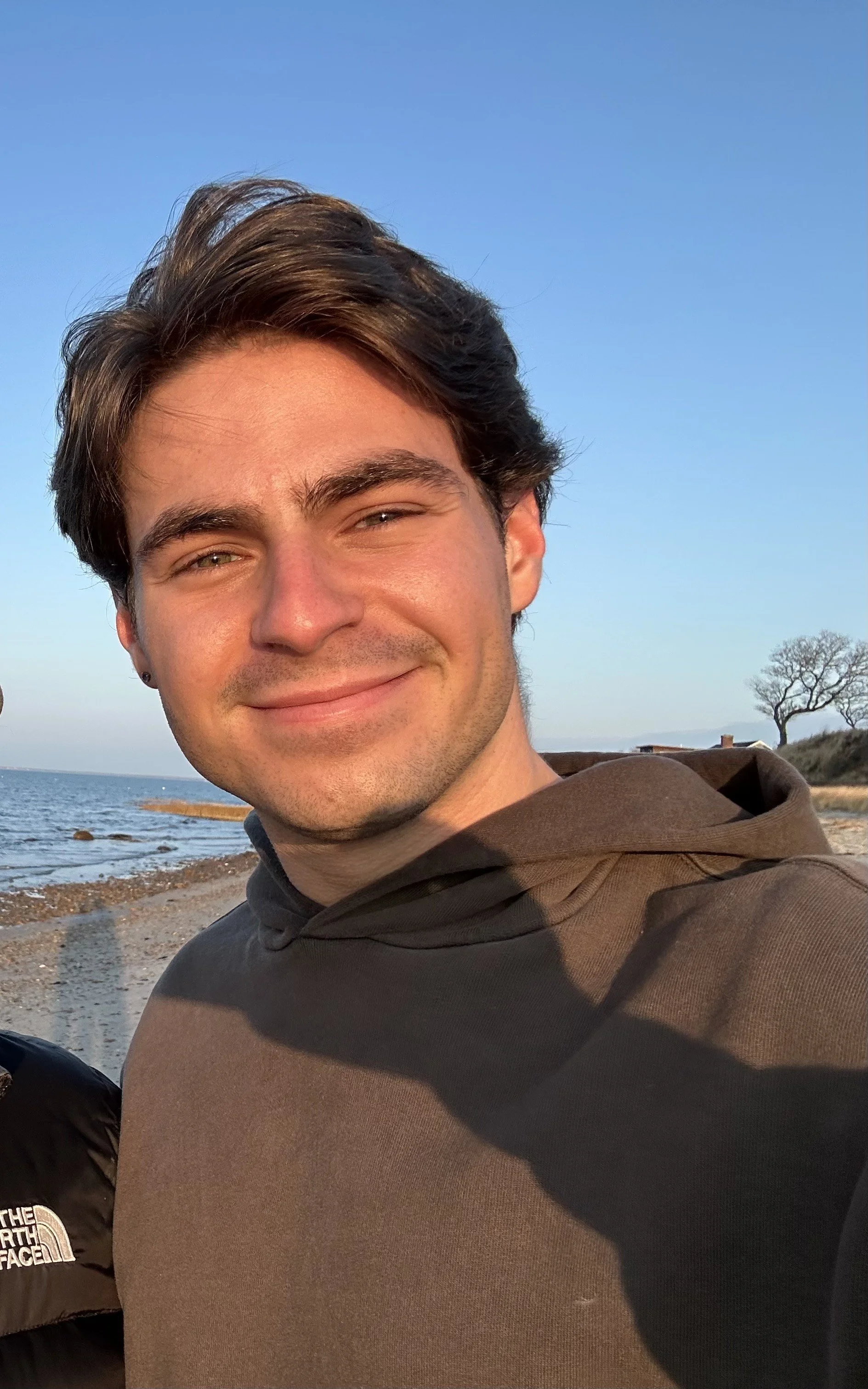 A young man with dark hair and a slight smile, wearing a brown hoodie, stands on a beach with the ocean and a leafless tree in the background during what appears to be late afternoon or early evening.