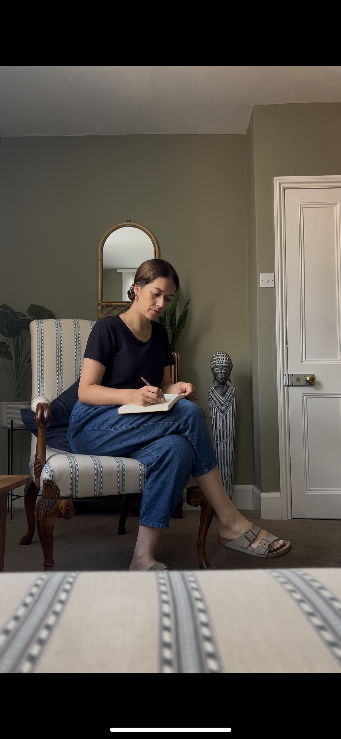A therapist sitting on a therapy chair, journaling in a notebook, in a therapy room with a green wall, a mirror, and decorative items, including a statue and plants.