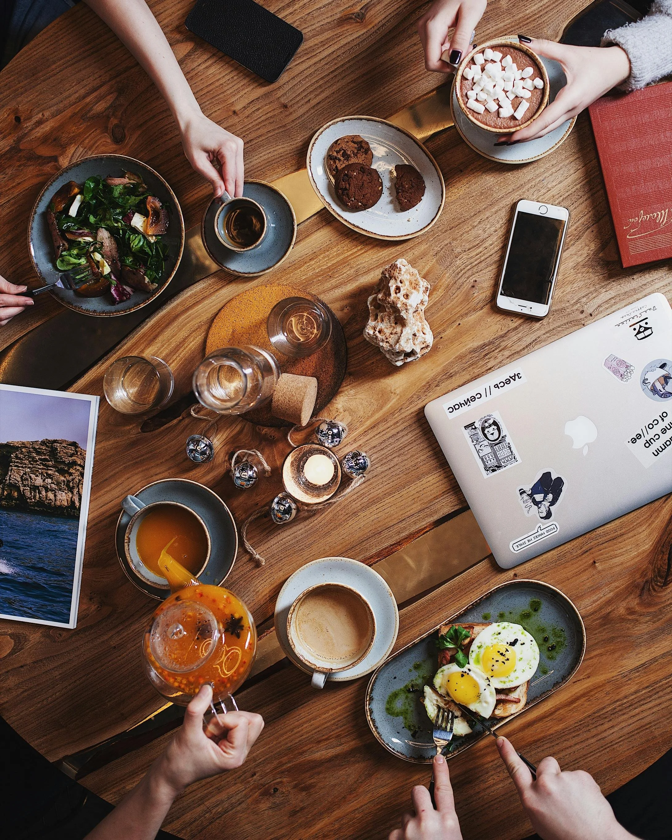 A top-down view of a wooden dining table with various foods, drinks, and personal items, including salad, cookies, a phone, coffee, mugs, a bowl of hot chocolate topped with marshmallows, an iced drink, and a laptop.