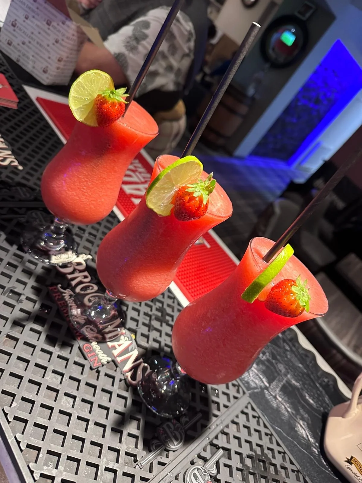 Three pink frozen strawberry cocktails garnished with lime slices and strawberries, with black straws, on a patterned table in a restaurant or bar setting.