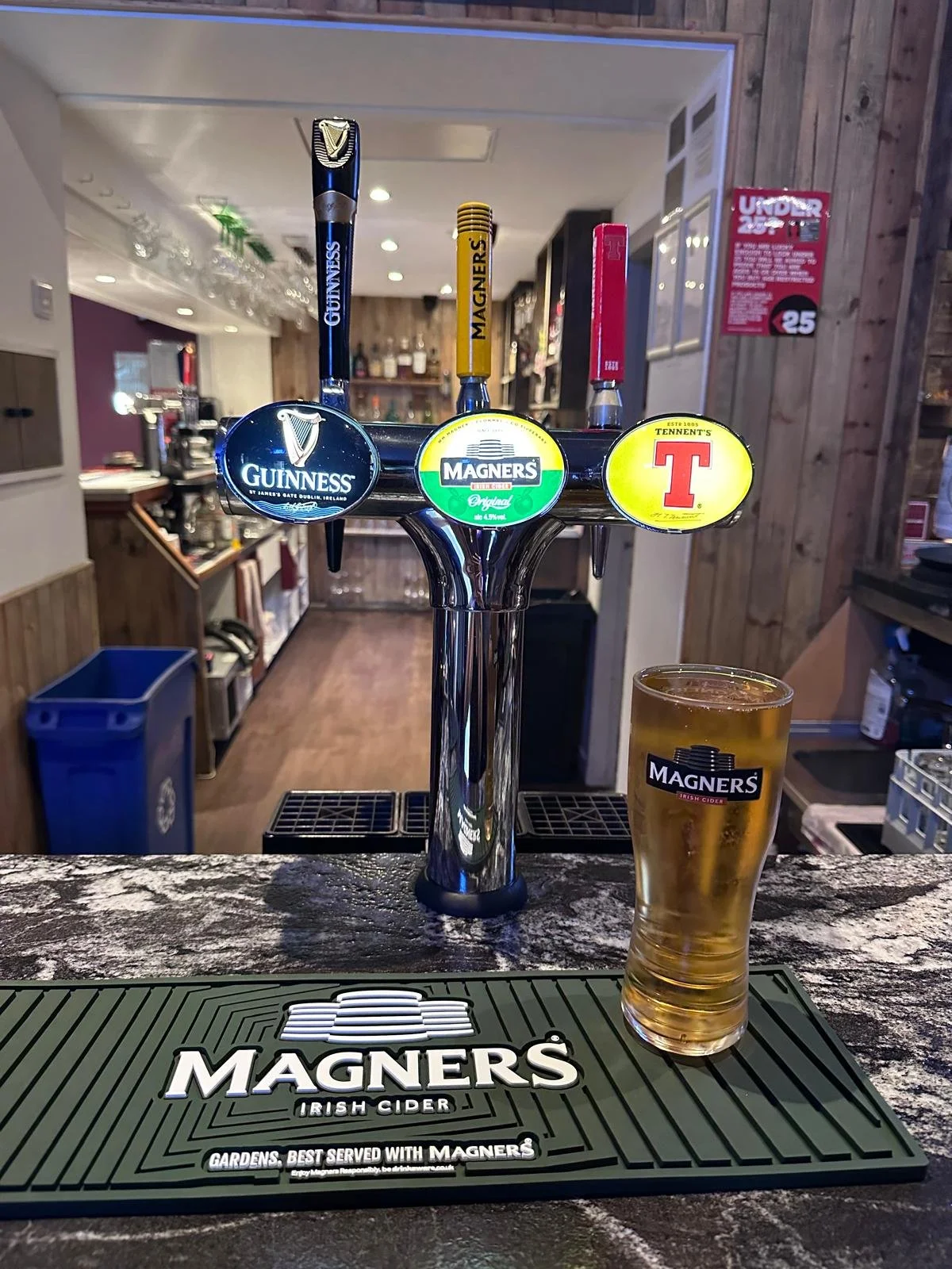 Bar with four beer tap handles labeled Guinness, Magners, and Tennent's, and a glass of Magners Irish cider on a bar mat.