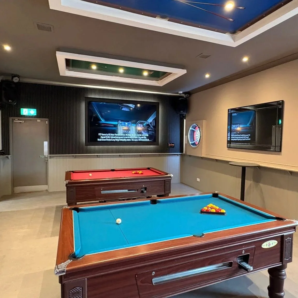 Pool game room with two pool tables, one with blue felt and one with red felt, and flat-screen televisions on the wall.