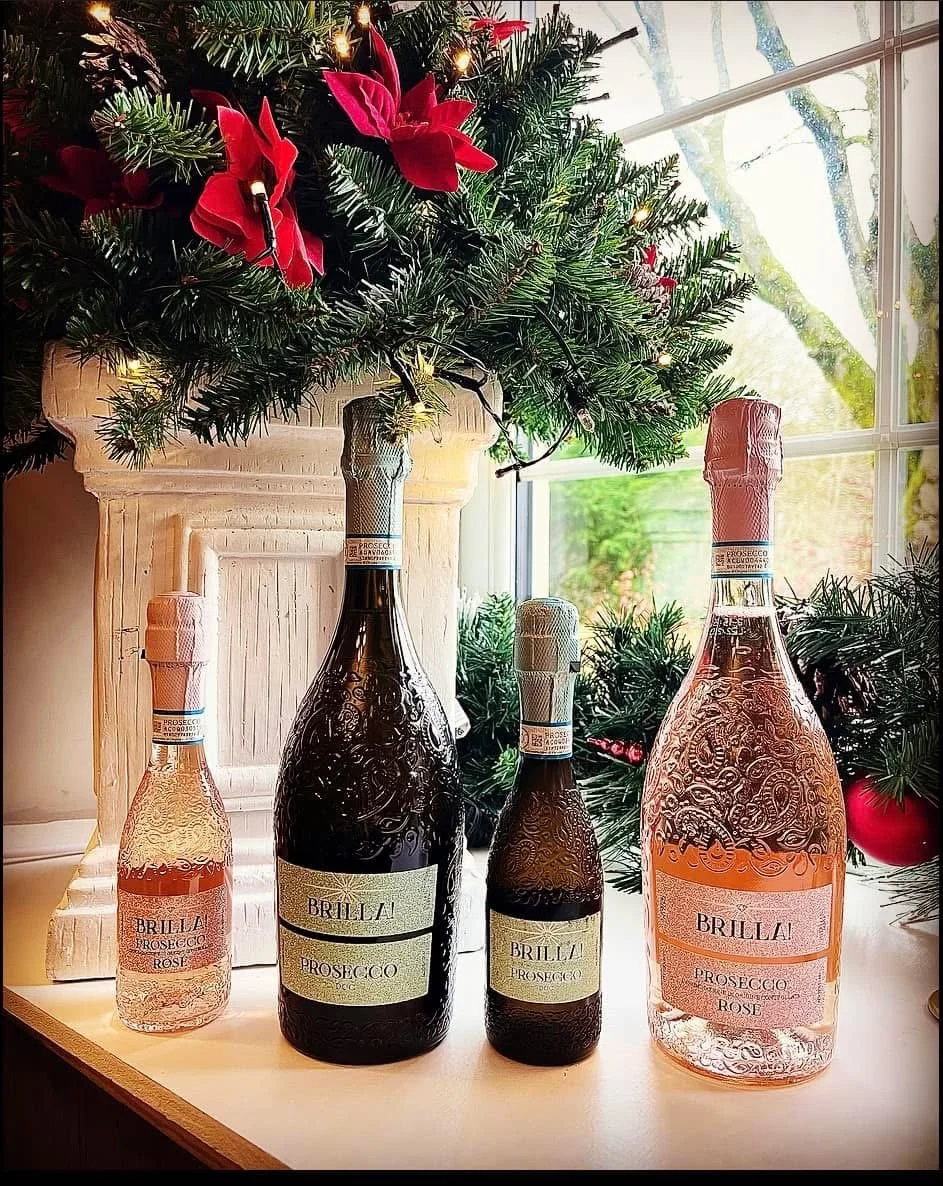Four bottles of Brilla Prosecco Rose in different sizes and a Christmas tree with red poinsettia decorations and lights on a windowsill.