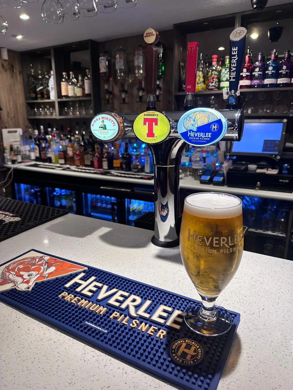 A pint of beer on a bar counter with a Welcome to Heverlee Premium Pilsner bar mat, behind it a beer tap with three handles labeled Birra Moretti, Tennent's, and Heverlee, and a range of liquor bottles on shelves in the background.