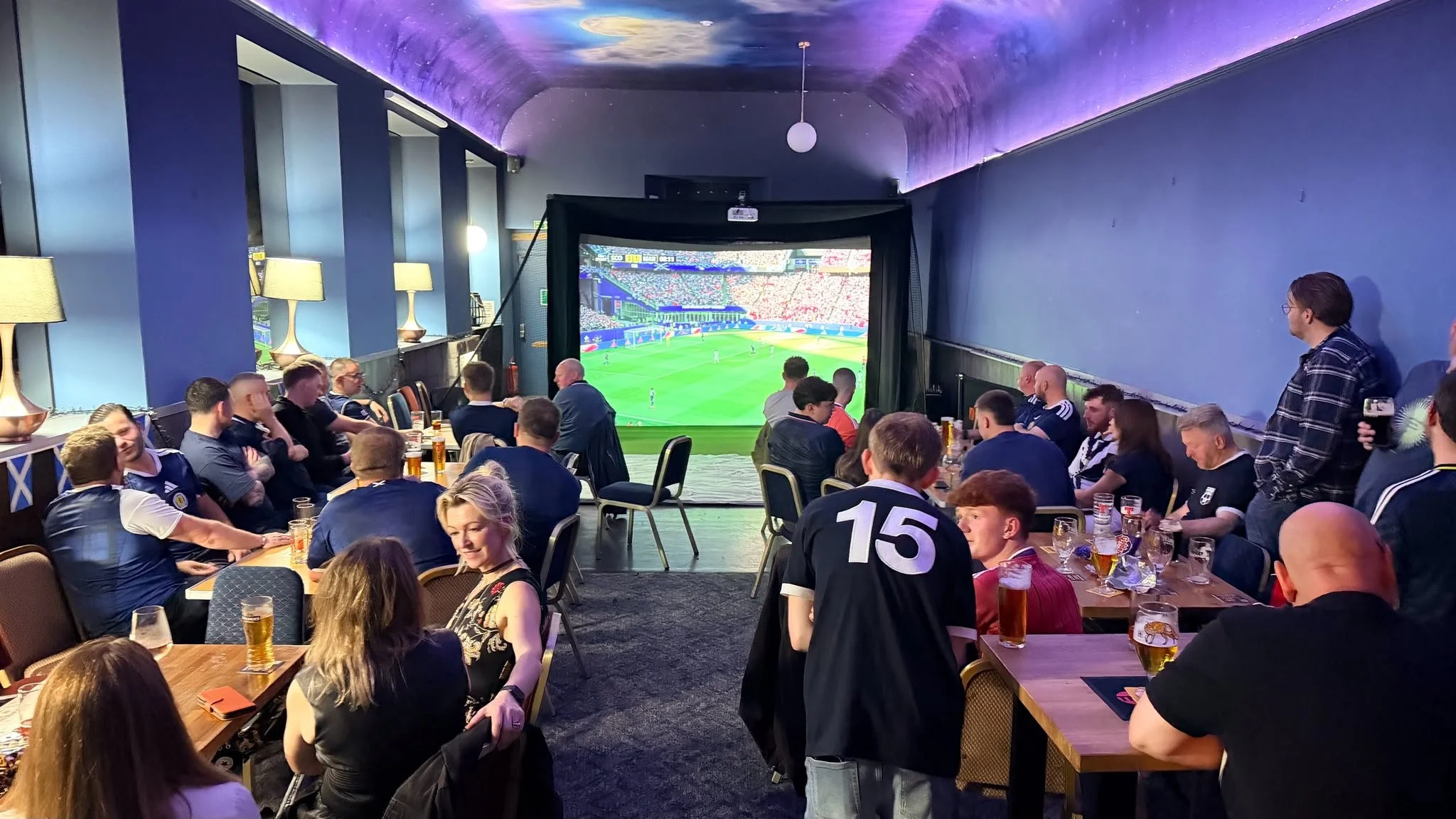 People sitting at tables in a room watching a large screen showing a soccer match. The room has a purple galaxy themed ceiling with purple lighting, and a backdrop with blue walls. Drinks are on the tables and some individuals are standing or talking.
