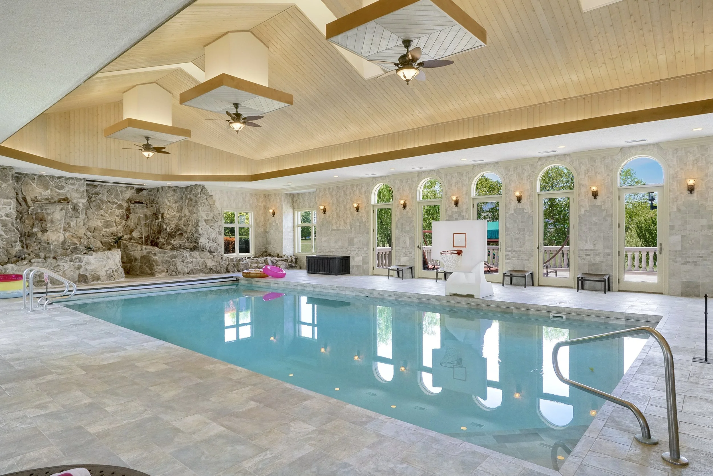 Indoor swimming pool with large windows, stone wall, and ceiling fans.